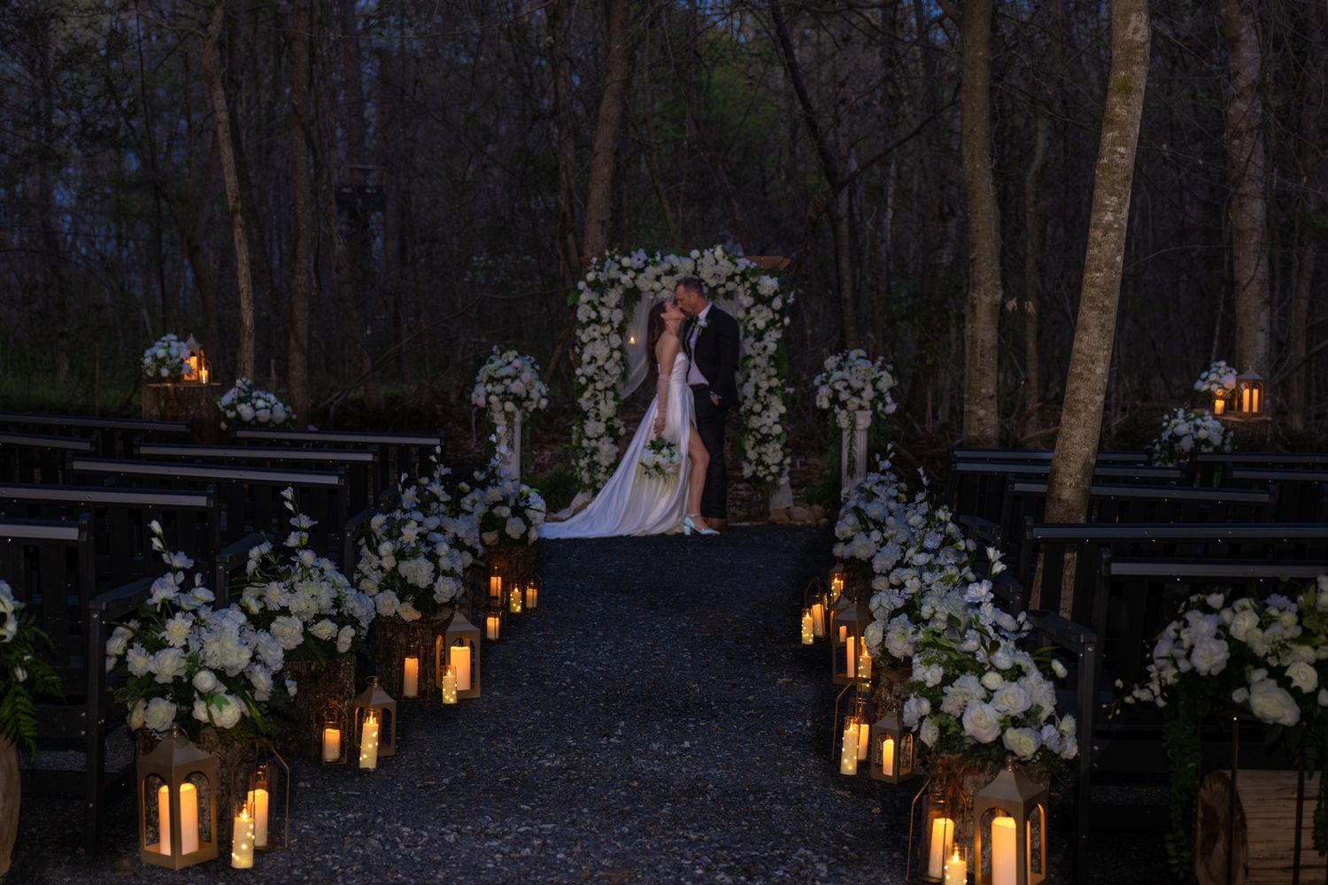 Romantic candlelit outdoor wedding ceremony at dusk with white floral arch, lanterns lining the aisle, and wooded backdrop.