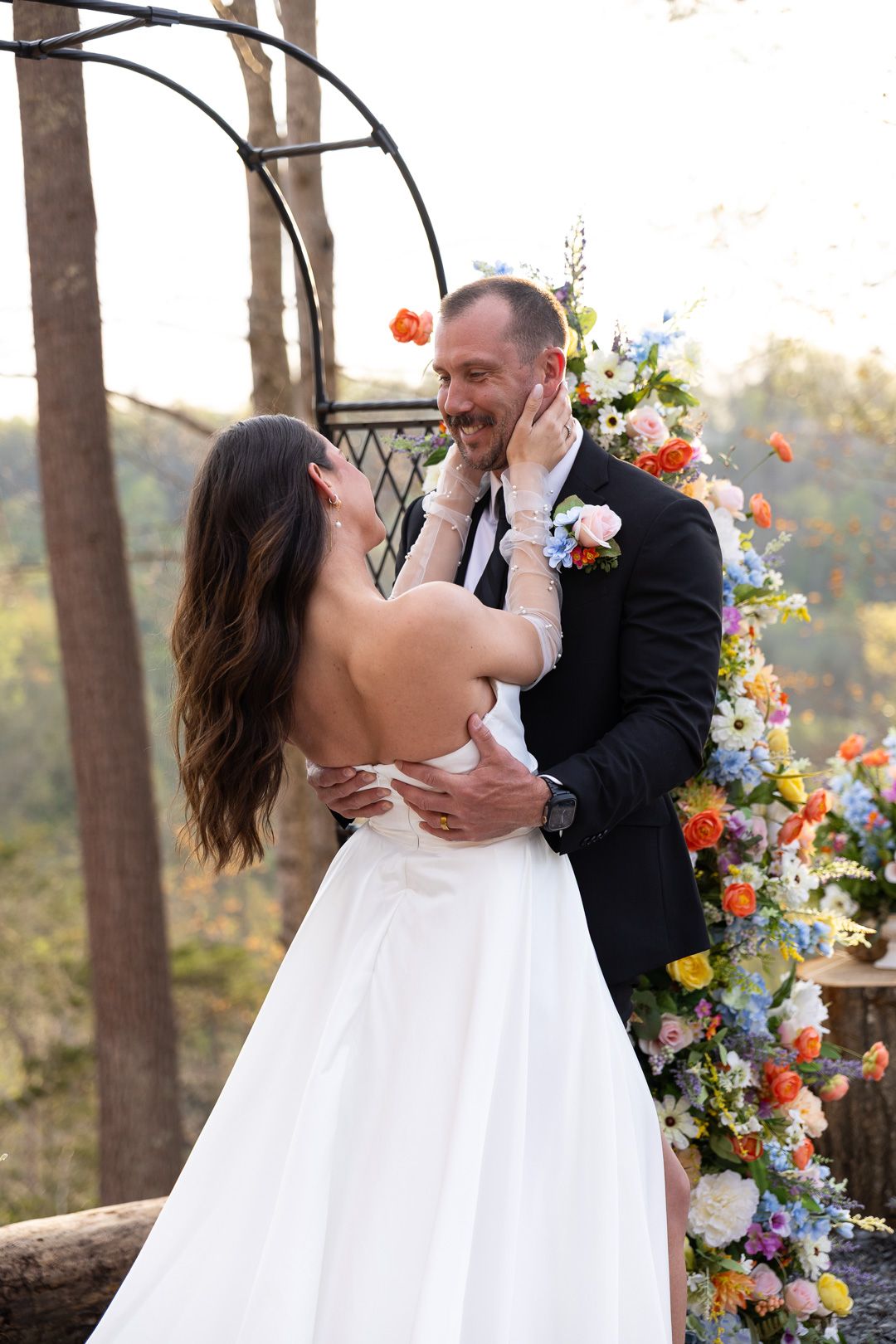 Bride tenderly cupping grooms smiling face at a colorful floral wedding arch ceremony in an outdoor woodland setting.