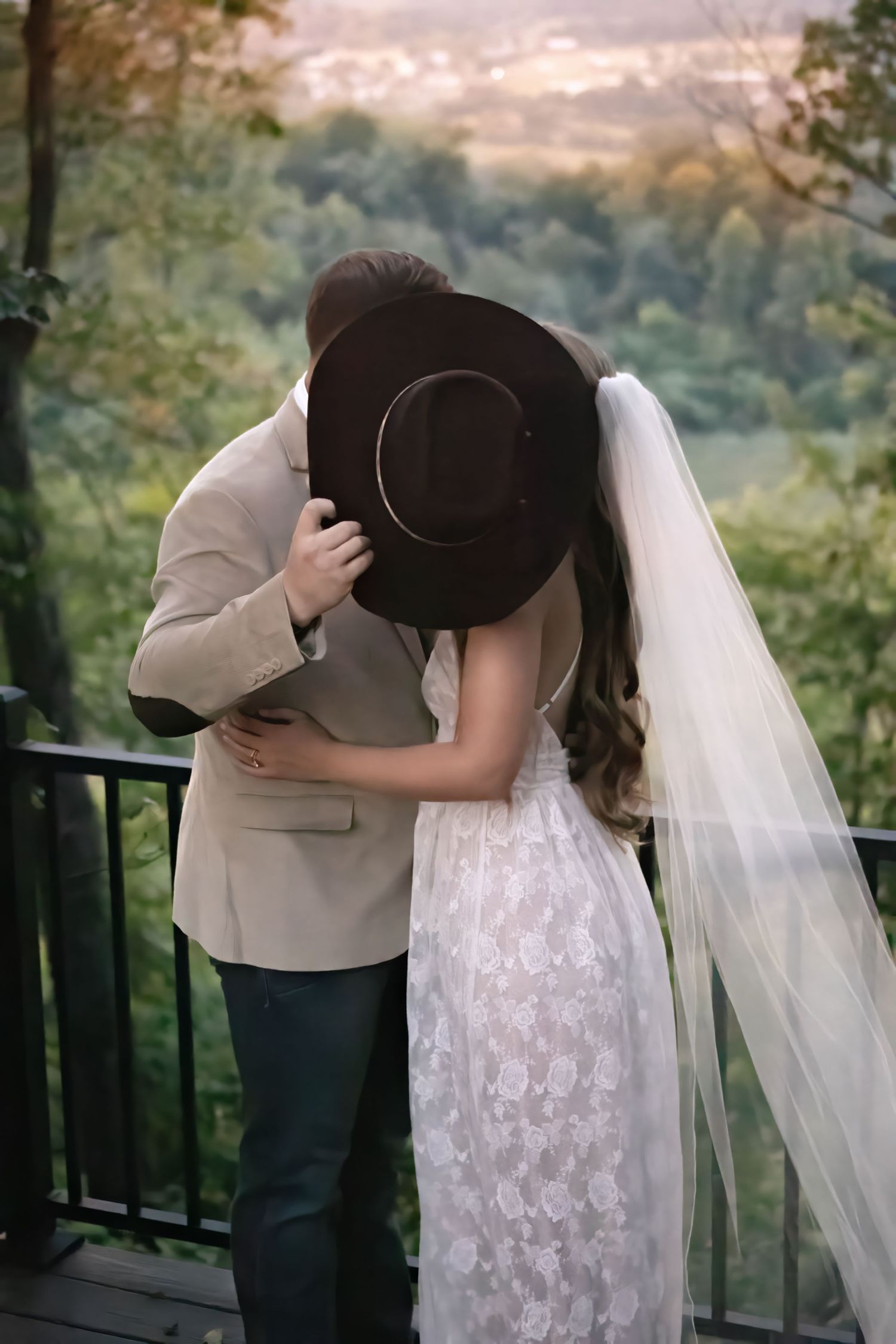 Bride and groom share a kiss hidden behind a cowboy hat on a scenic overlook, bride wearing lace dress and veil.