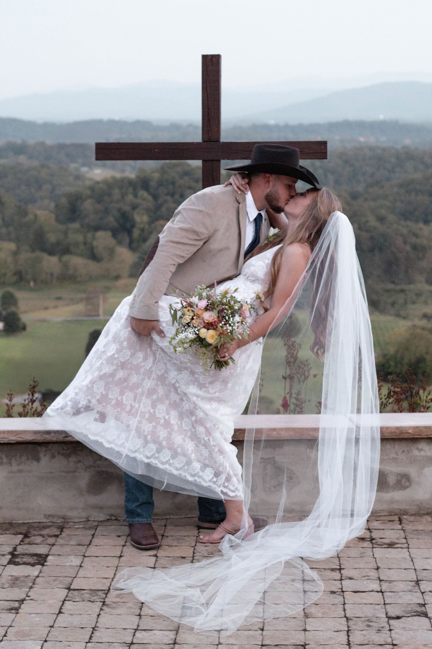 Bride and groom share a romantic kiss in front of a wooden cross with scenic mountain views on their wedding day.