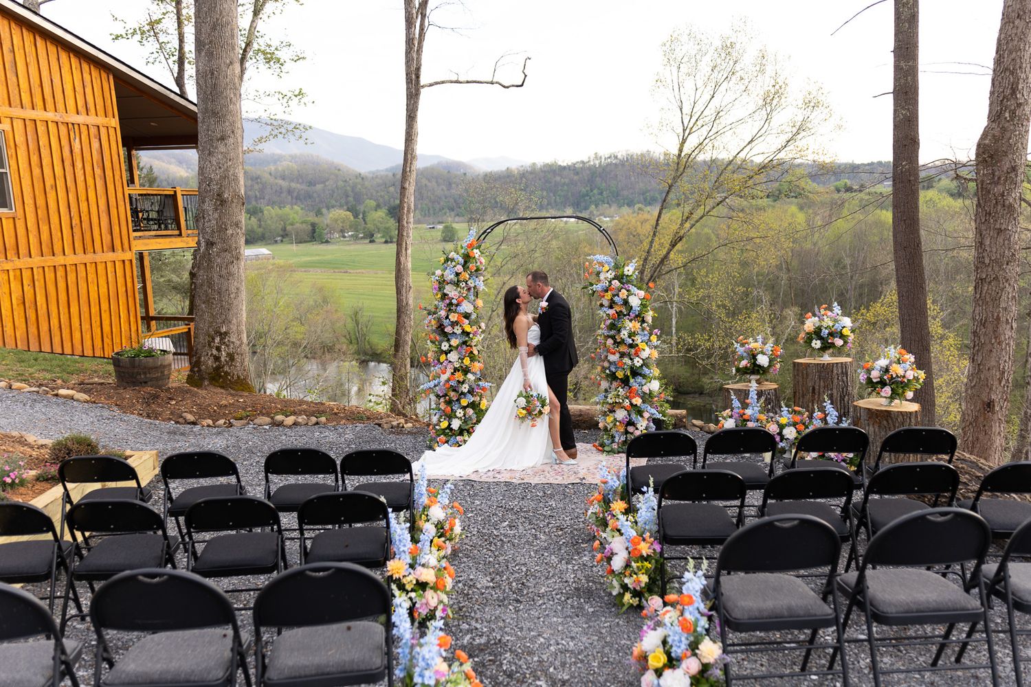 Outdoor wedding ceremony setup with colorful floral arch, black chairs, and scenic mountain valley backdrop.