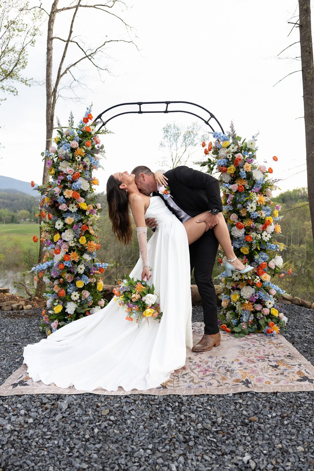 Bride and groom sharing a romantic dip kiss in front of a vibrant multicolored floral wedding ceremony arch.