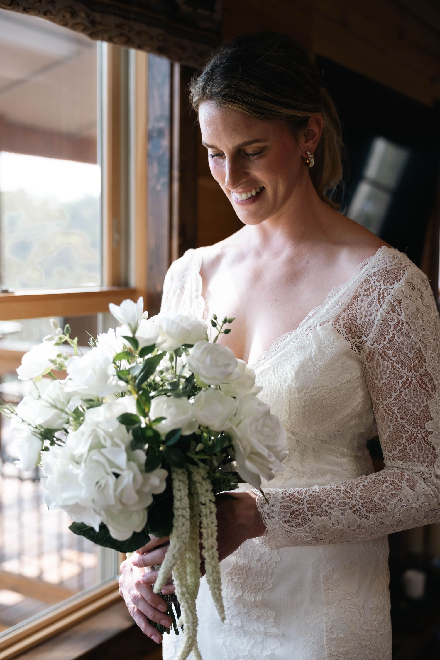 Smiling bride in lace long-sleeve wedding dress holding white rose bouquet near window.