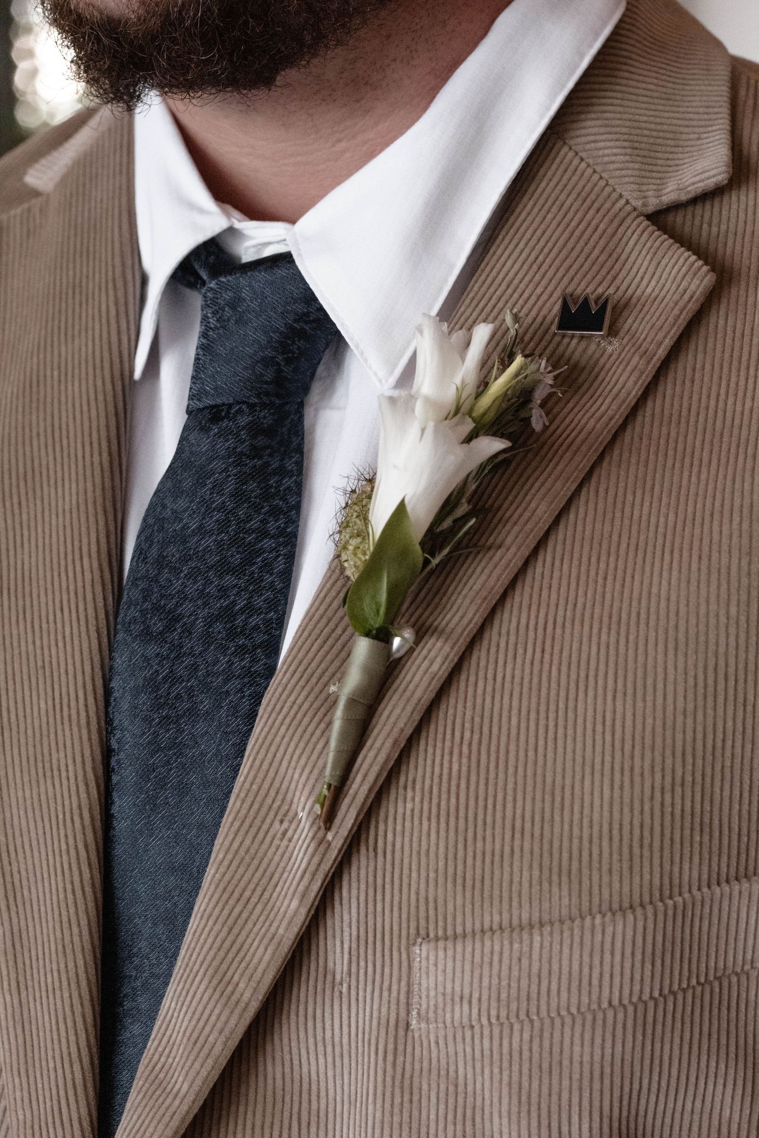 Groom wearing tan corduroy suit with dark floral tie and white boutonniere with greenery pinned to lapel.