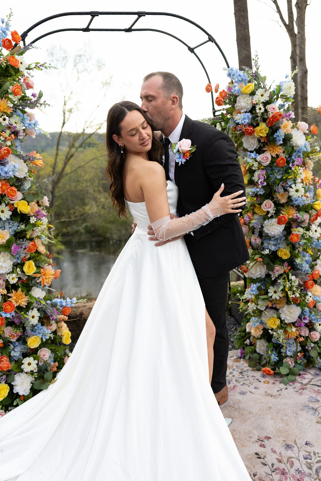 Bride and groom kissing under a colorful floral arch with river backdrop on their wedding day.