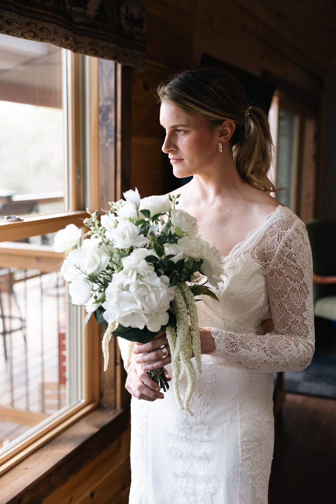 Bride in lace long-sleeve wedding dress holds white floral bouquet while gazing out a rustic cabin window.