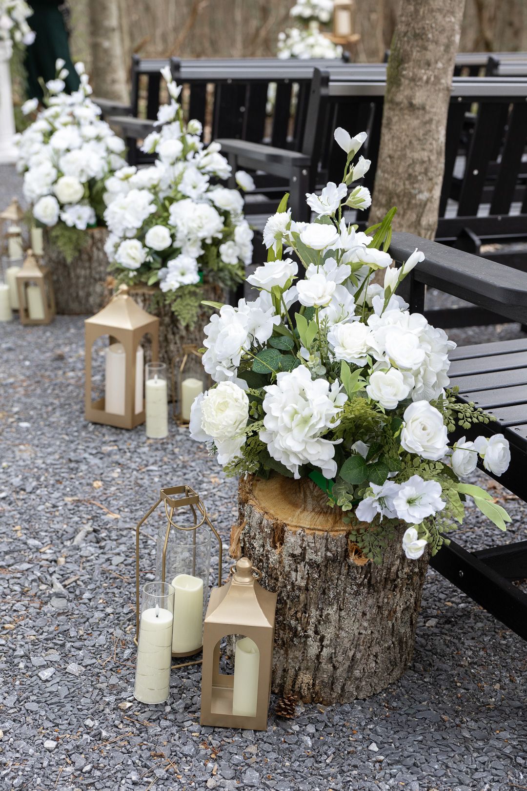 White floral arrangements on tree stumps with gold lanterns and candles lining a rustic outdoor wedding aisle.