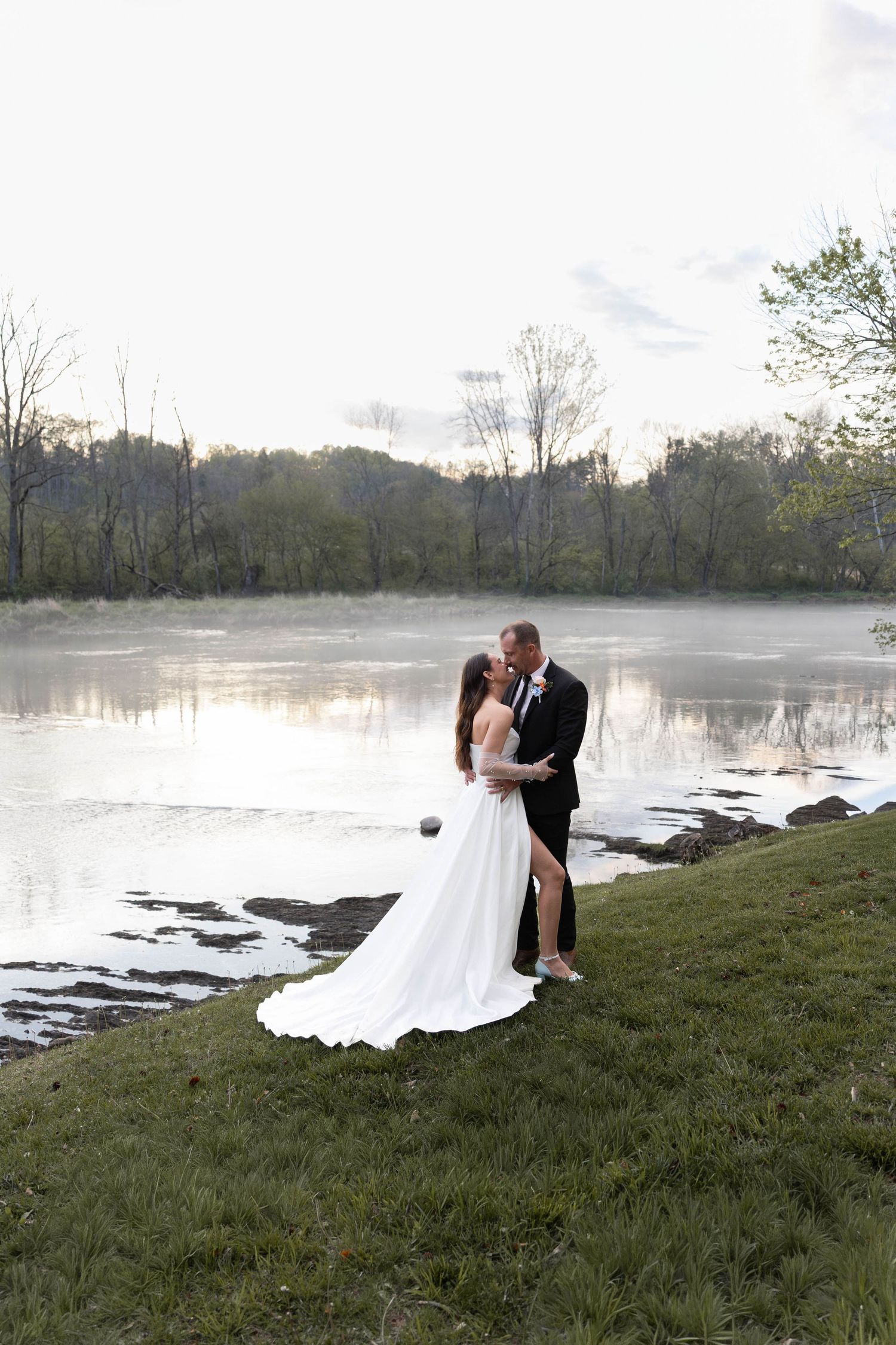 Bride and groom share a romantic kiss by a misty river at sunset, surrounded by lush greenery in a serene outdoor setting.
