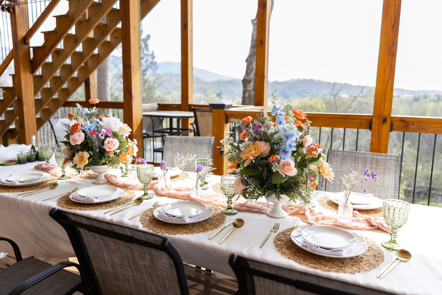 Elegant outdoor wedding reception table set with floral centerpieces, gold chargers, and colorful blooms on a wooden deck.