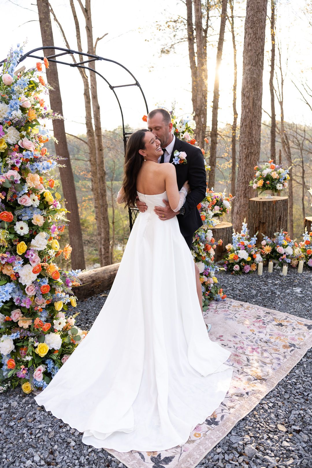 Bride and groom embrace at outdoor wedding altar adorned with colorful flowers and arch, surrounded by trees at sunset.