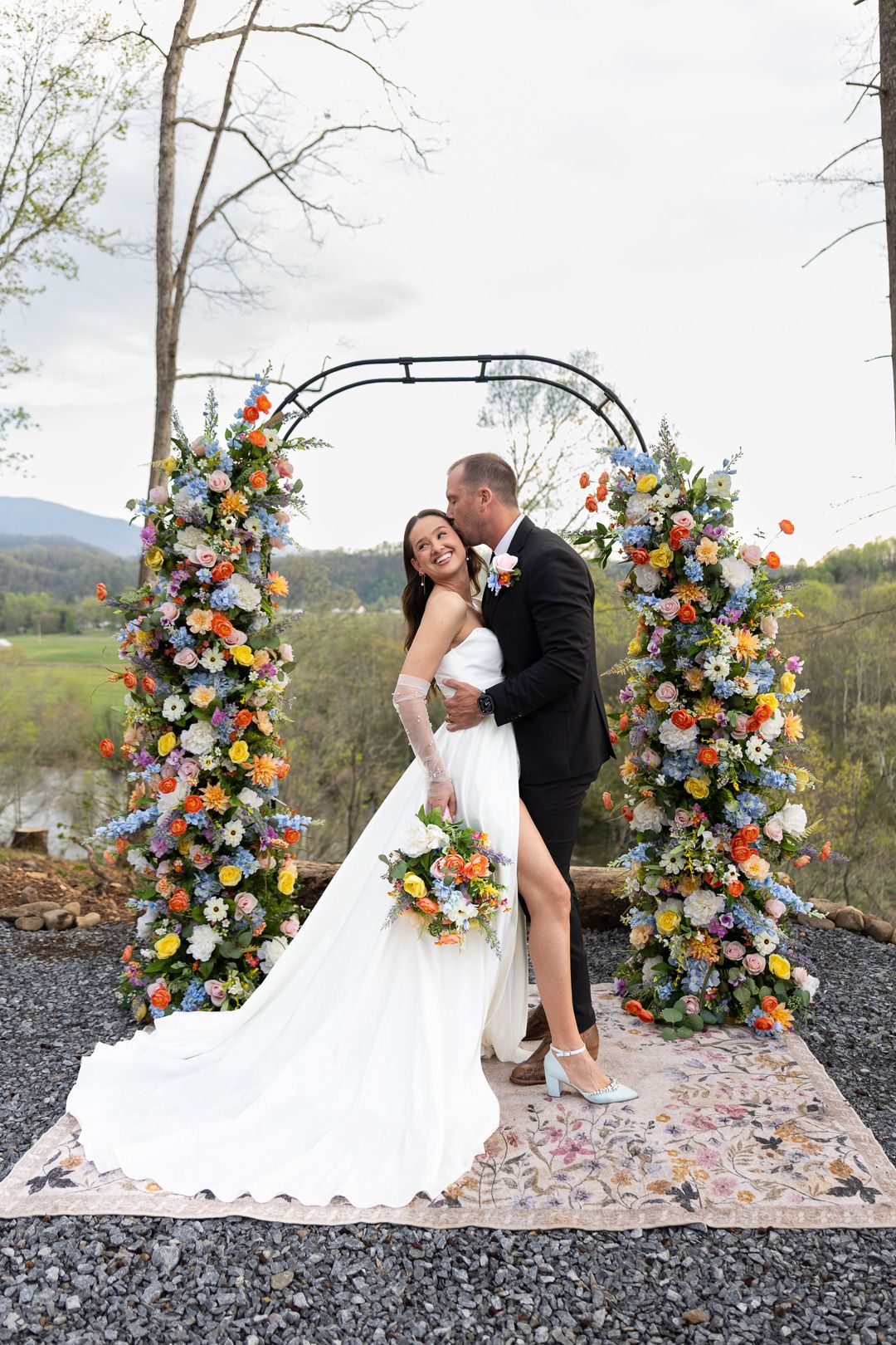 Bride and groom embrace under a colorful floral arch at an outdoor wedding ceremony with mountain views.