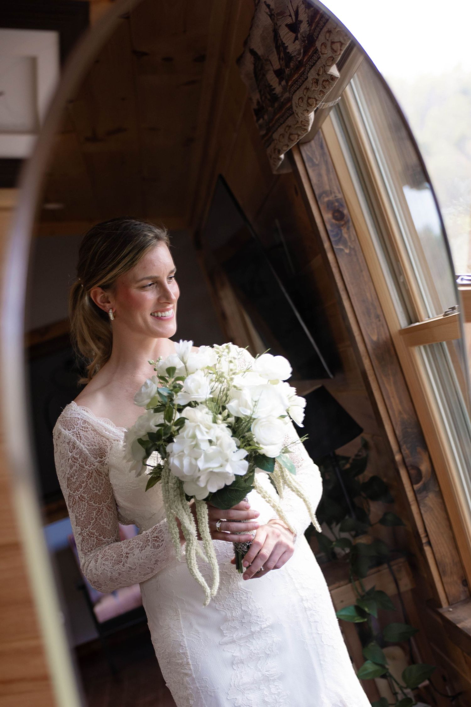 Smiling bride in lace wedding dress holding white hydrangea bouquet standing by wooden arch window.