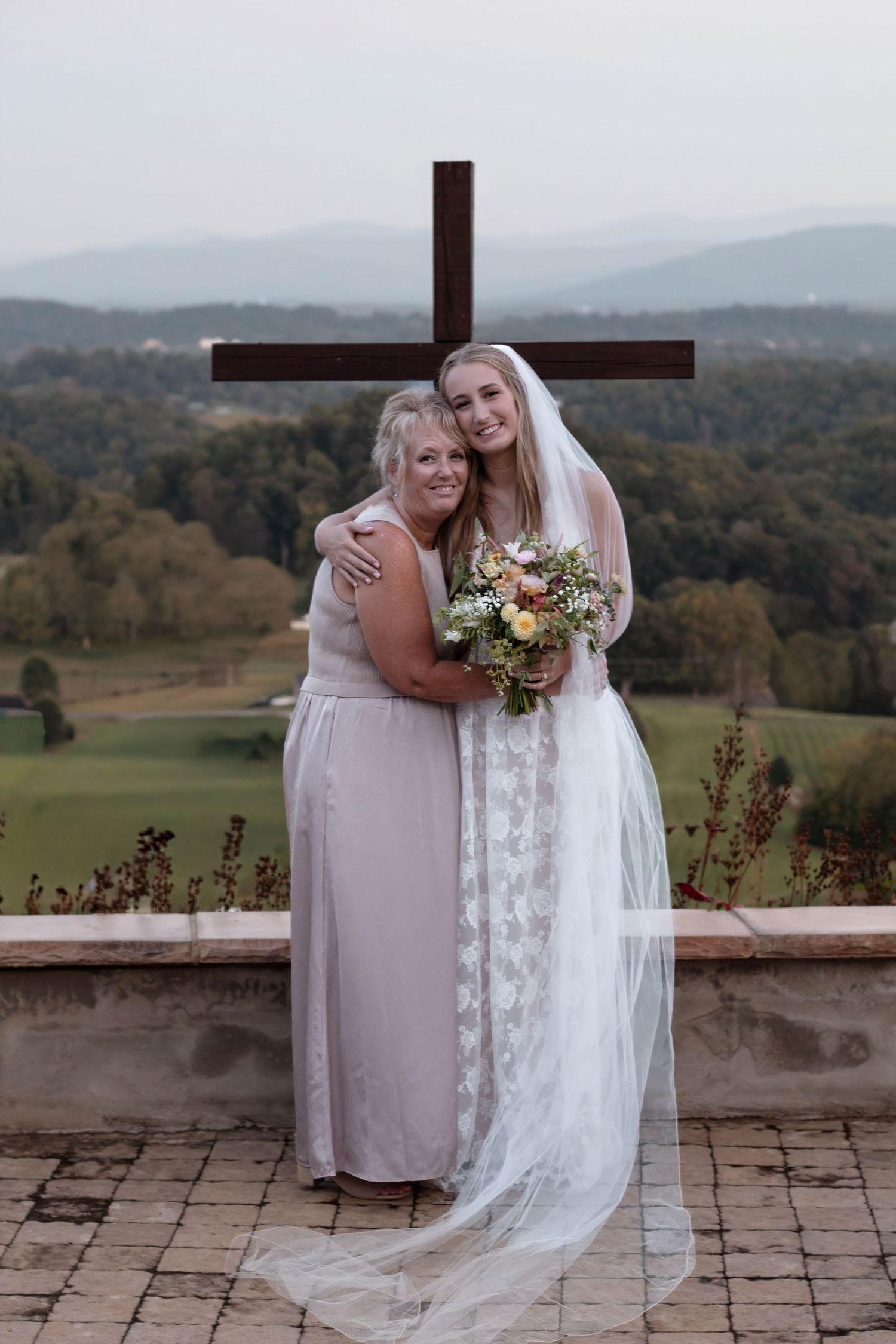 Bride in lace gown and veil holds bouquet beside bridesmaid in front of wooden cross with scenic mountain view.