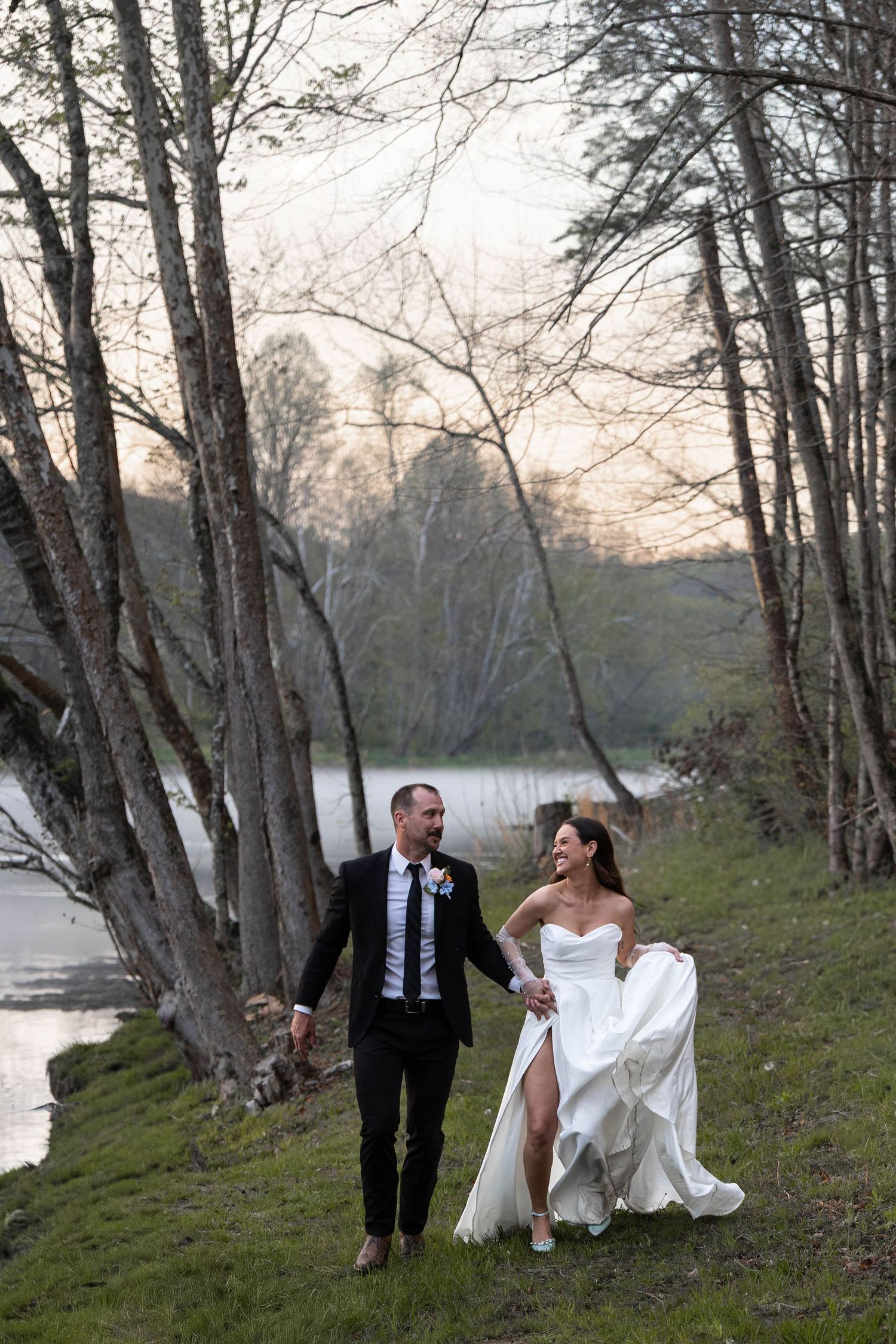 Newlyweds walking hand-in-hand along a riverside path at sunset, bride lifting her white gown, groom in black suit.
