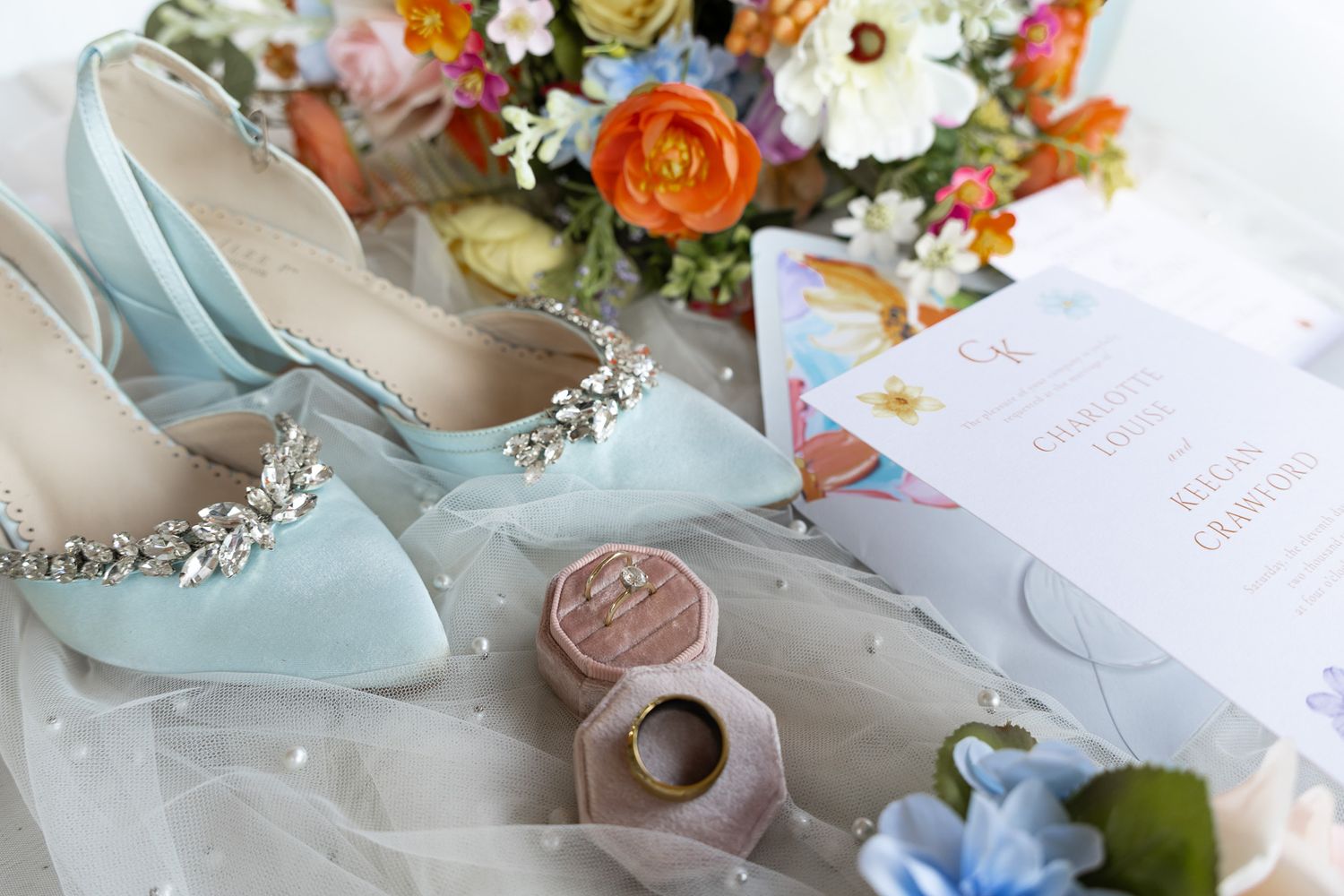 Light blue bridal heels with crystal straps beside a floral bouquet, ring box, and wedding invitation.