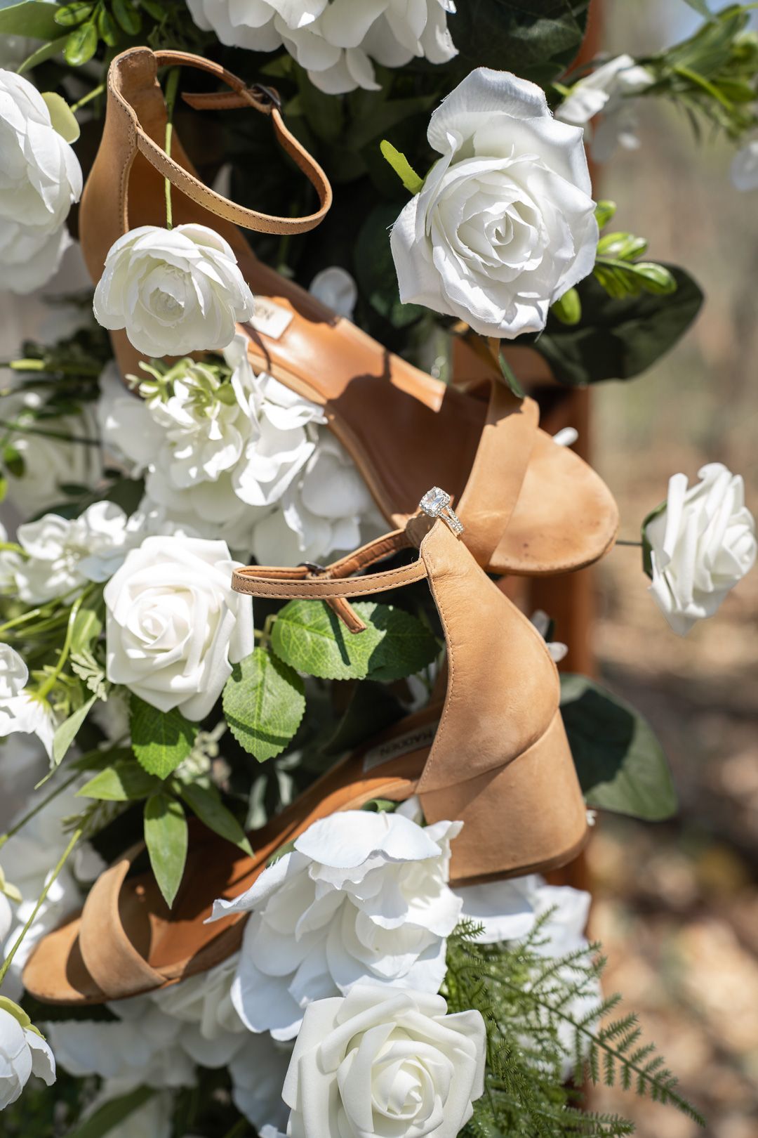 Tan suede bridal heels with ankle straps displayed among white roses and green foliage for a wedding.