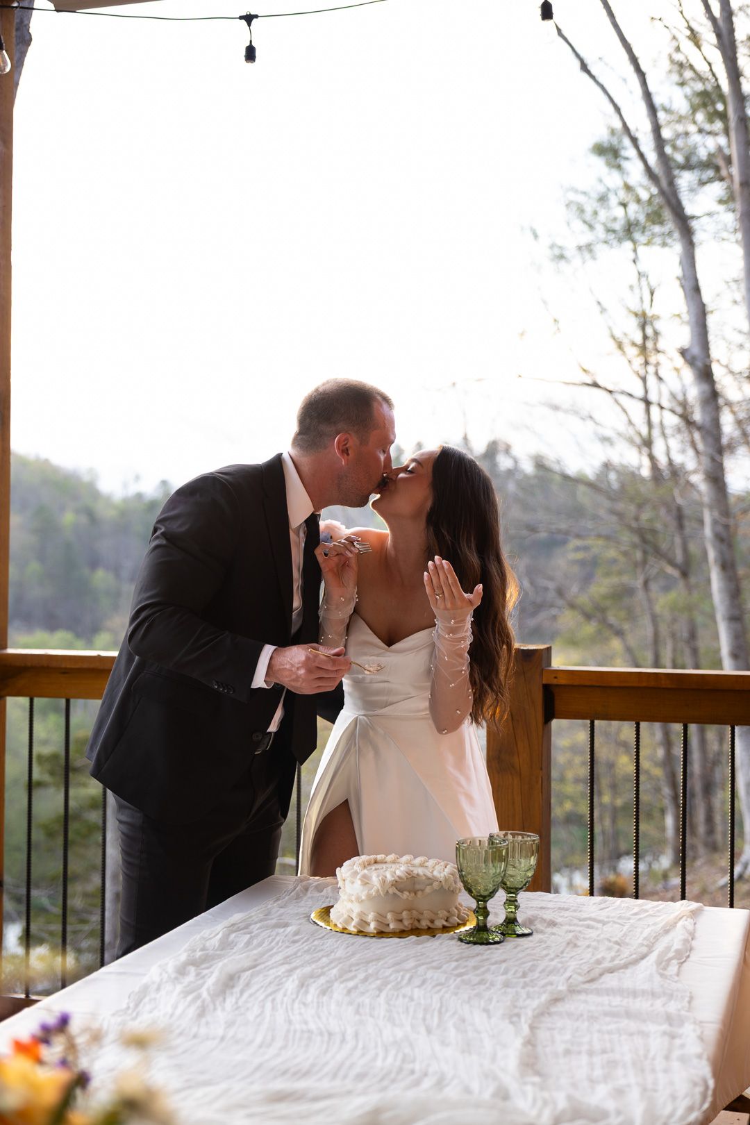 Bride and groom kissing while cutting their white wedding cake on a sunlit wooden deck with green goblets.
