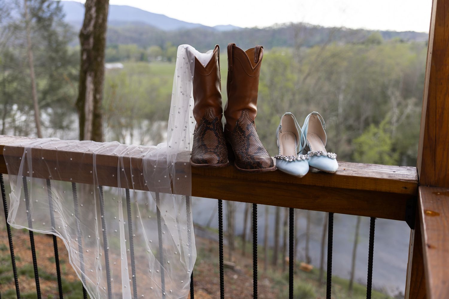 Cowboy boots and blue heels on a wooden porch railing with a lace veil and scenic view.