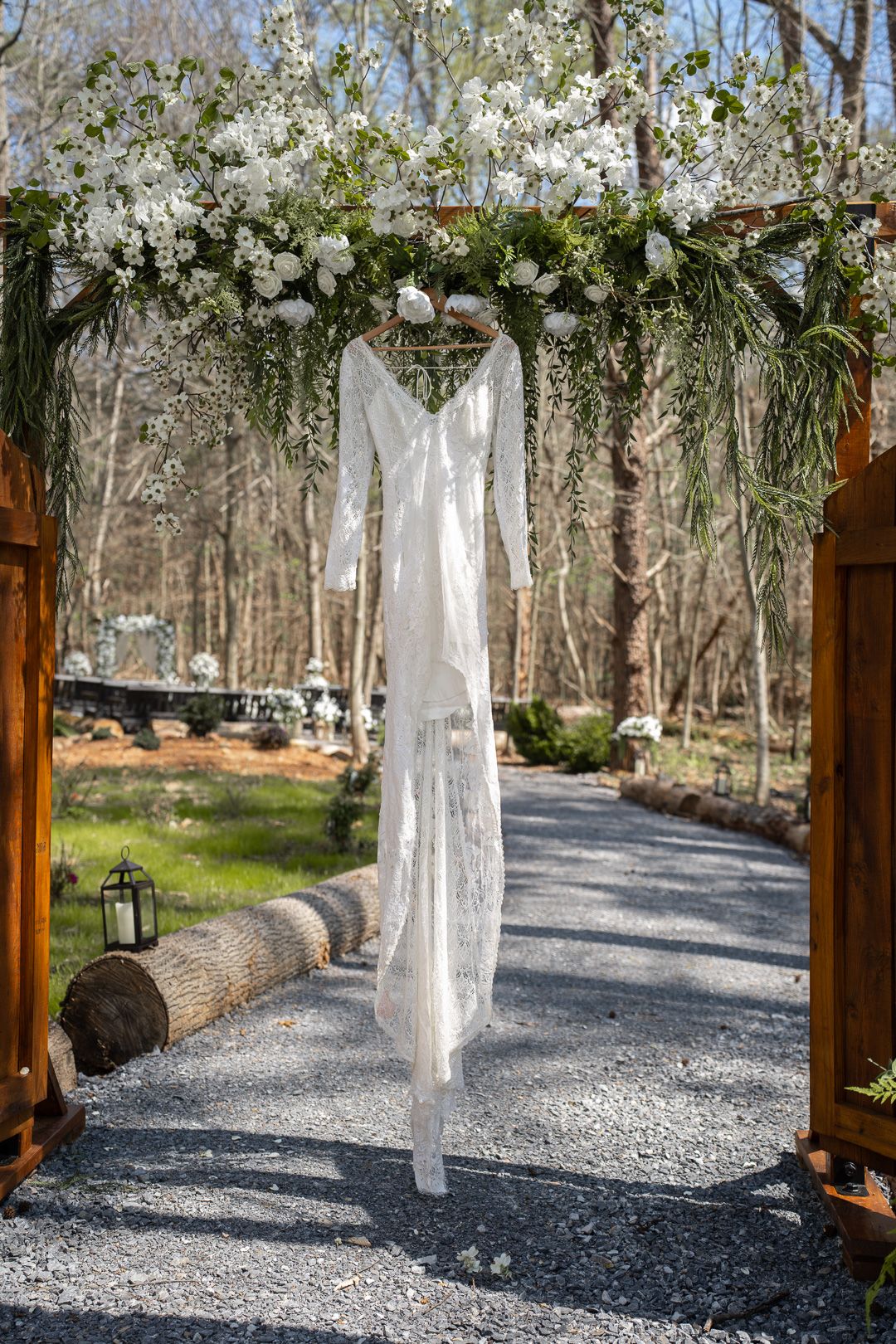 White lace wedding dress hanging from a floral arch at an outdoor woodland ceremony venue.