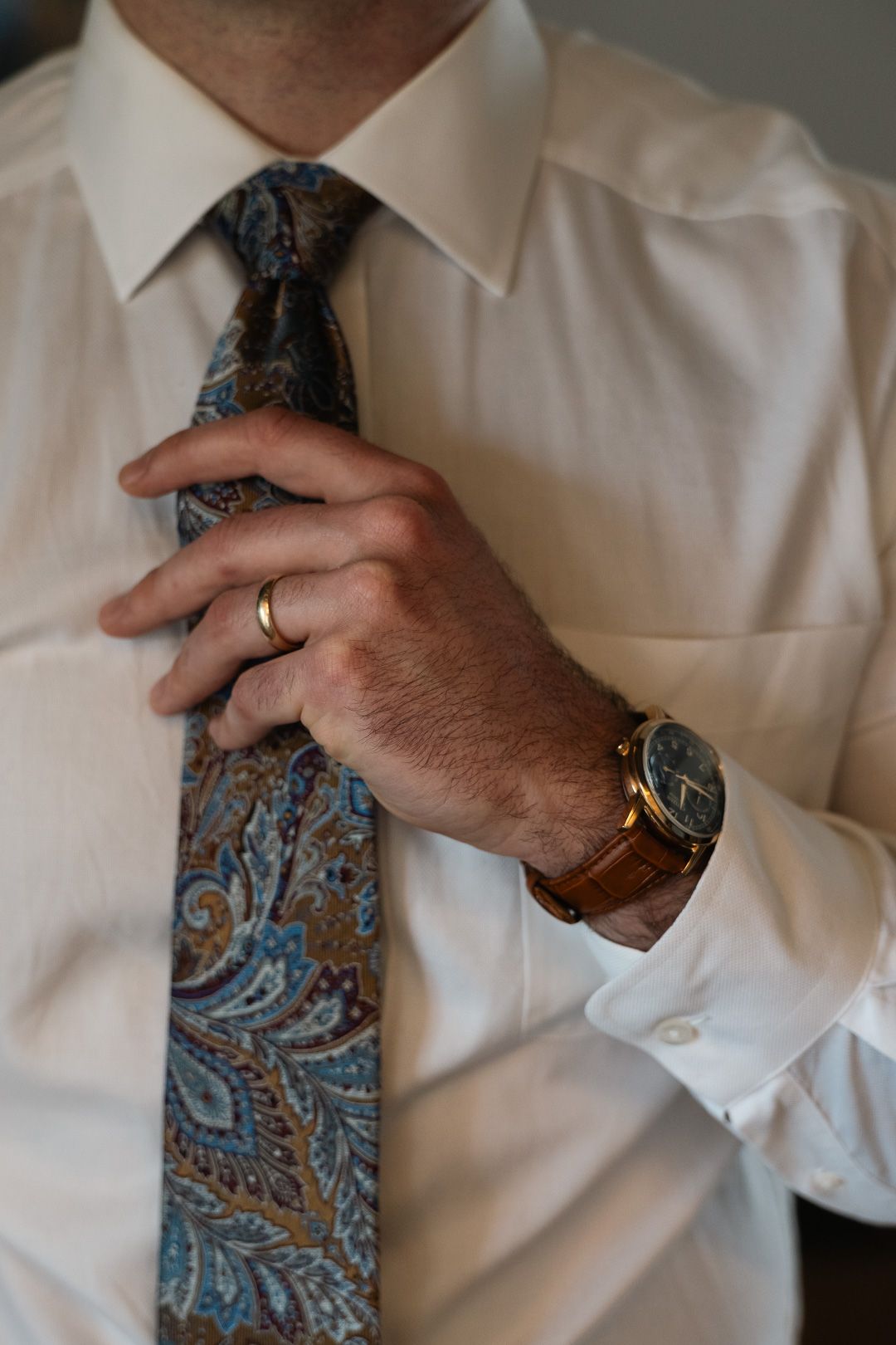 Man in white dress shirt adjusting paisley tie, wearing gold ring and brown leather strap watch.