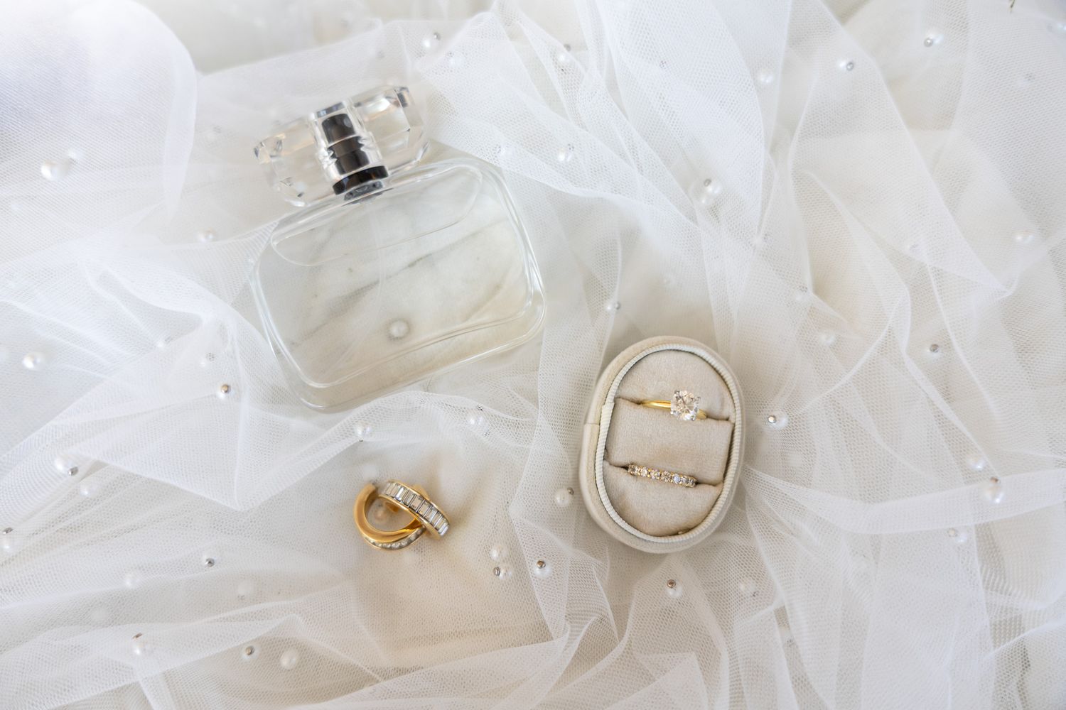 Wedding flat lay featuring a perfume bottle, diamond engagement ring, gold band, and pearl-adorned veil on white tulle.