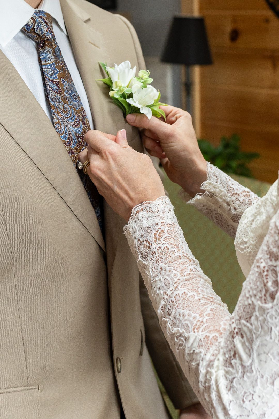 Bride pinning a white floral boutonniere on groom's beige suit lapel with paisley tie at wedding.