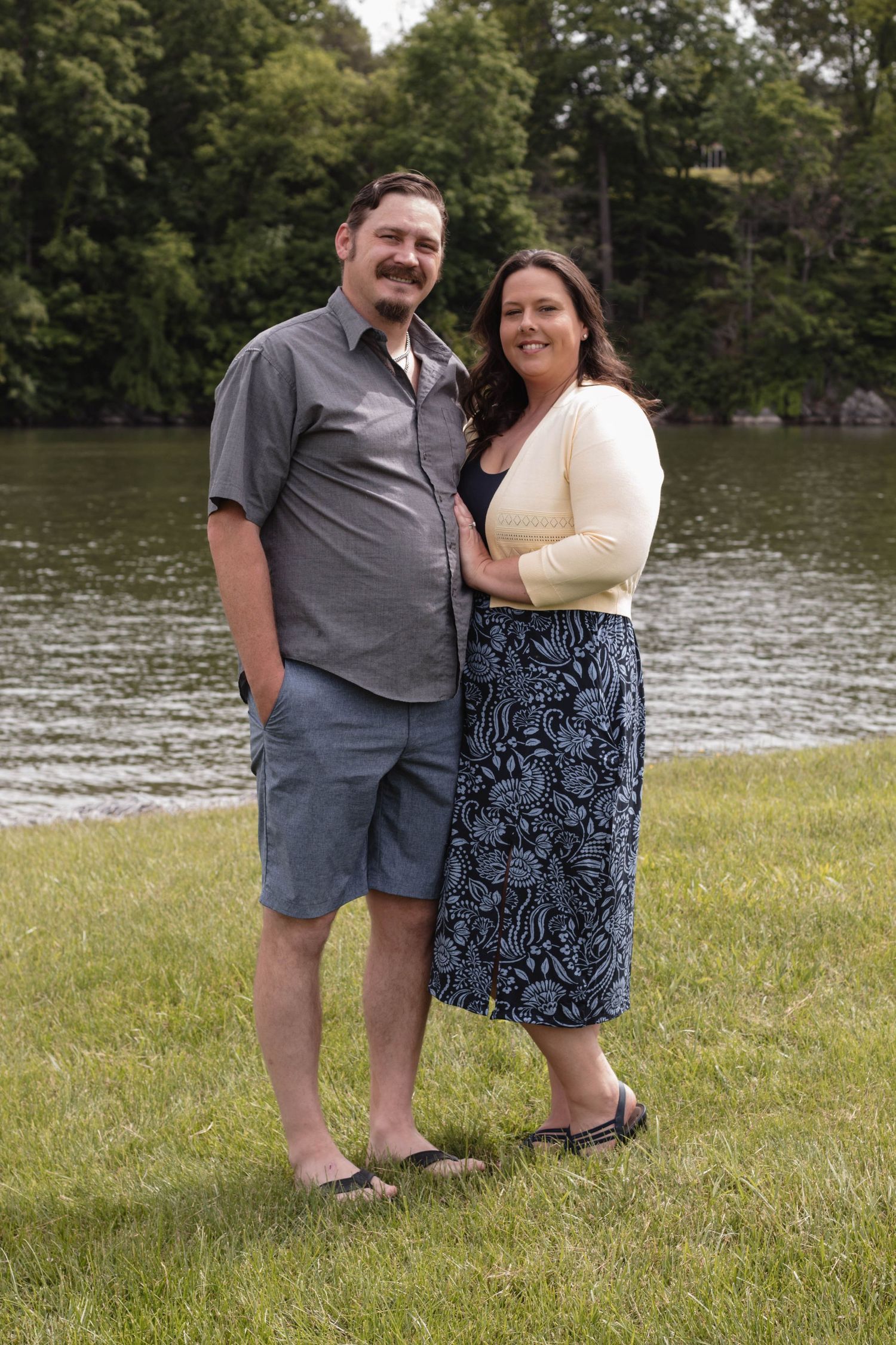 A couple stands together on grassy lawn by a lake wearing casual summer attire on a sunny day.