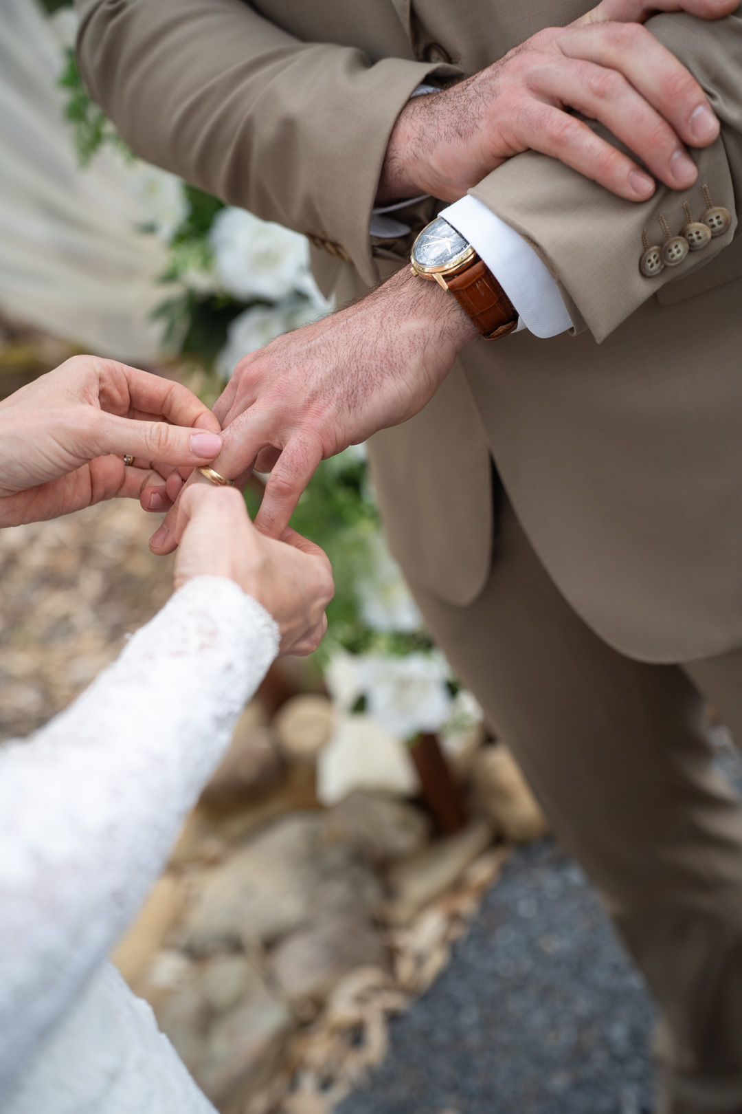 Bride placing a wedding ring on groom's finger during outdoor ceremony, groom wearing beige suit and gold watch.