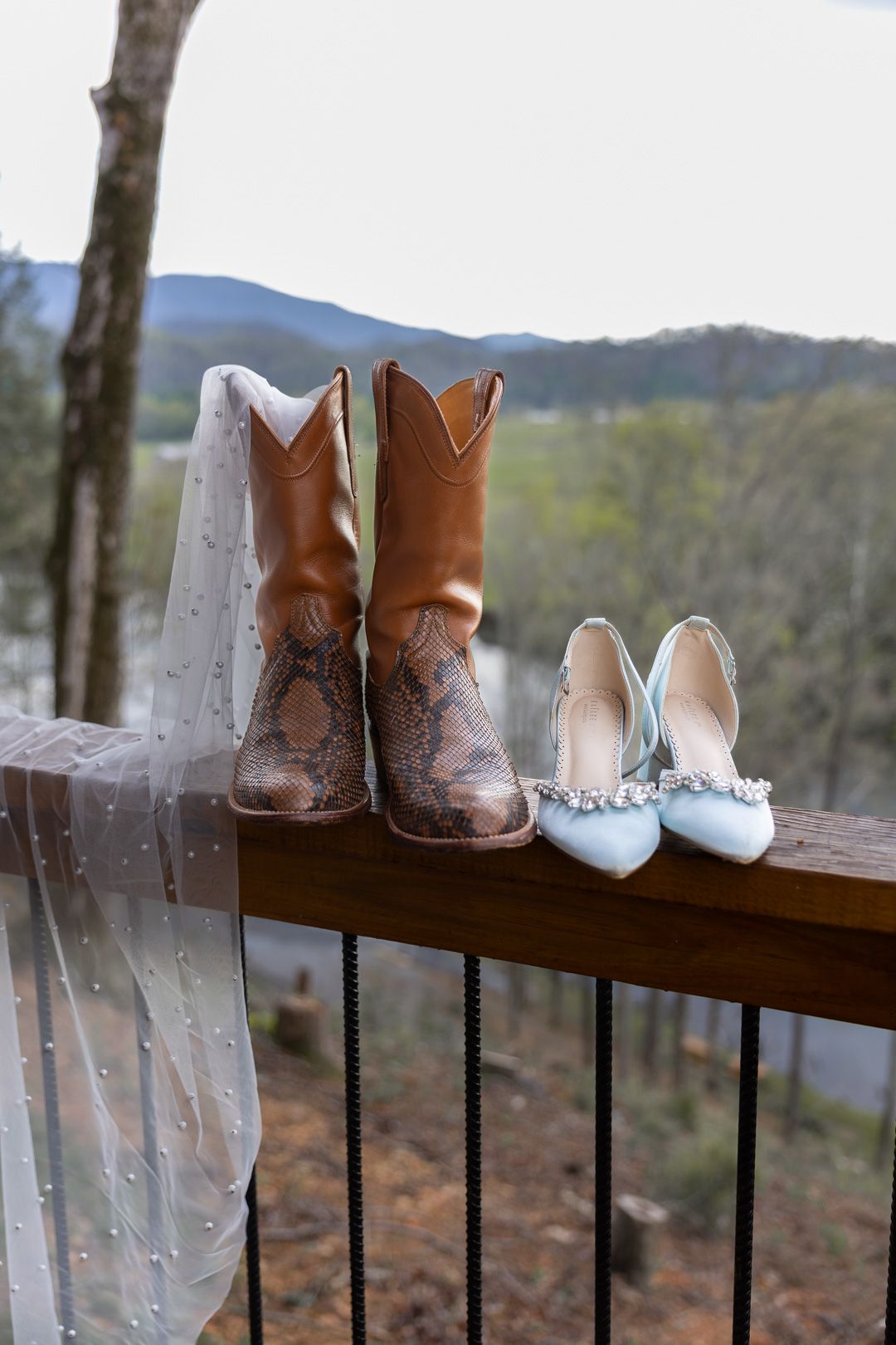 Bride's snakeskin cowboy boots and blue jeweled heels displayed on a wooden deck railing with a dotted veil.