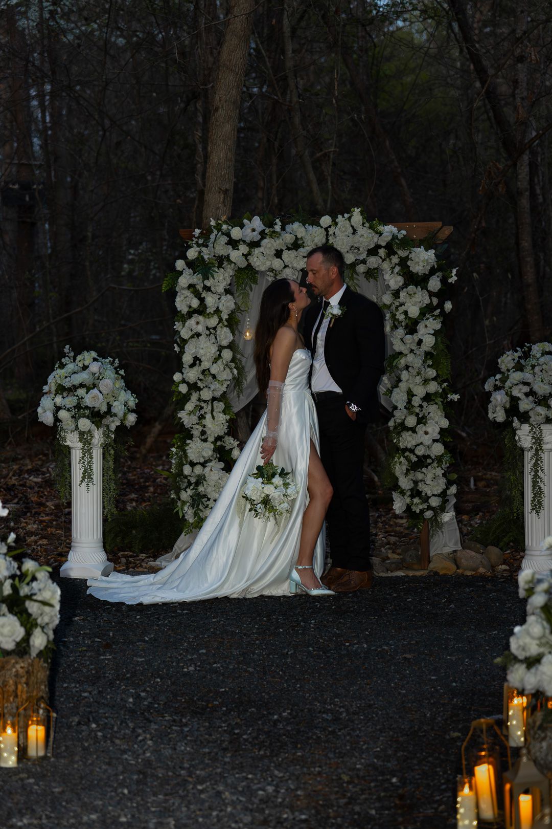 Bride and groom kissing under a white floral arch at dusk, flanked by candle lanterns and white flower columns.