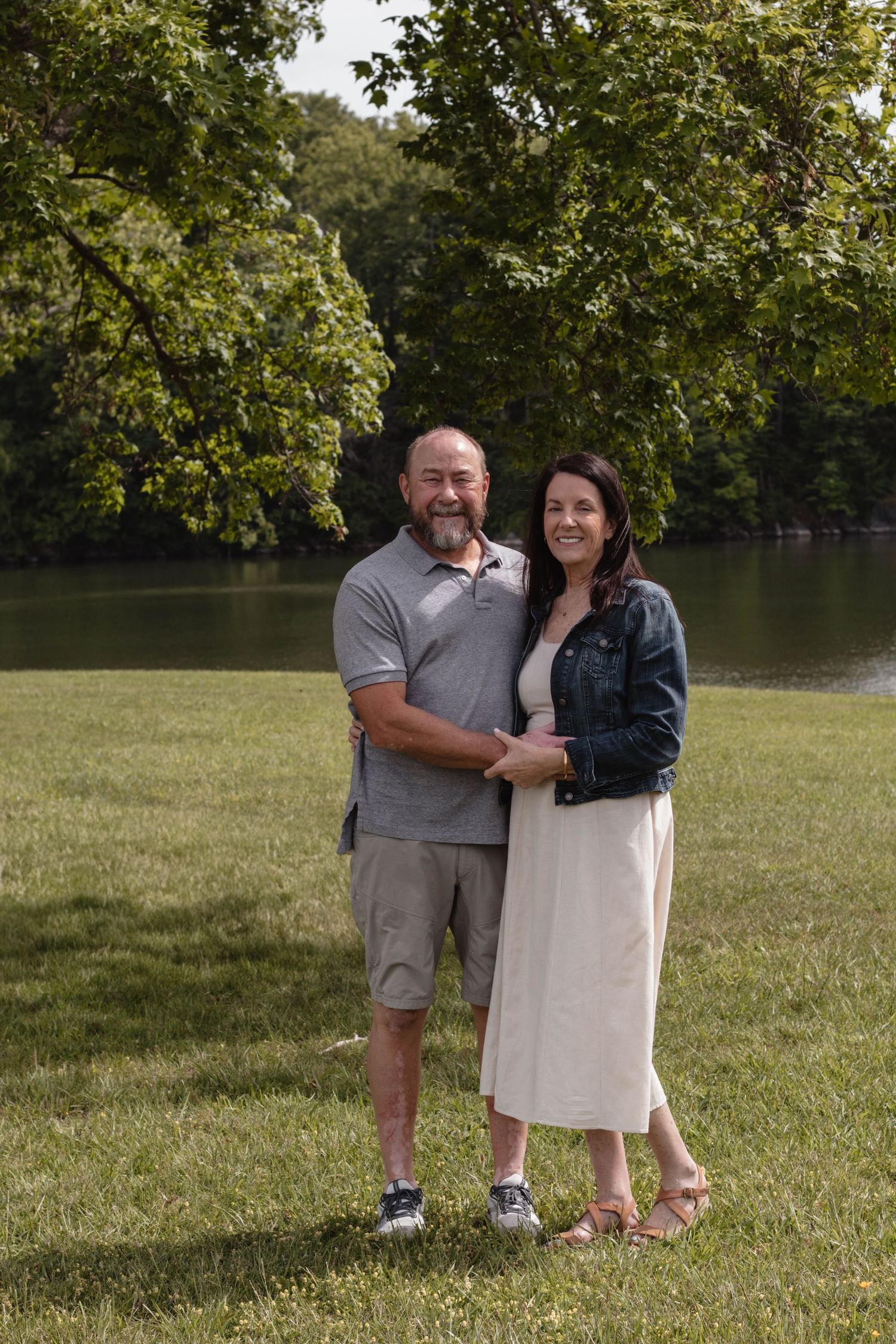 A couple embraces while standing on green grass with trees in the background during a sunny day.