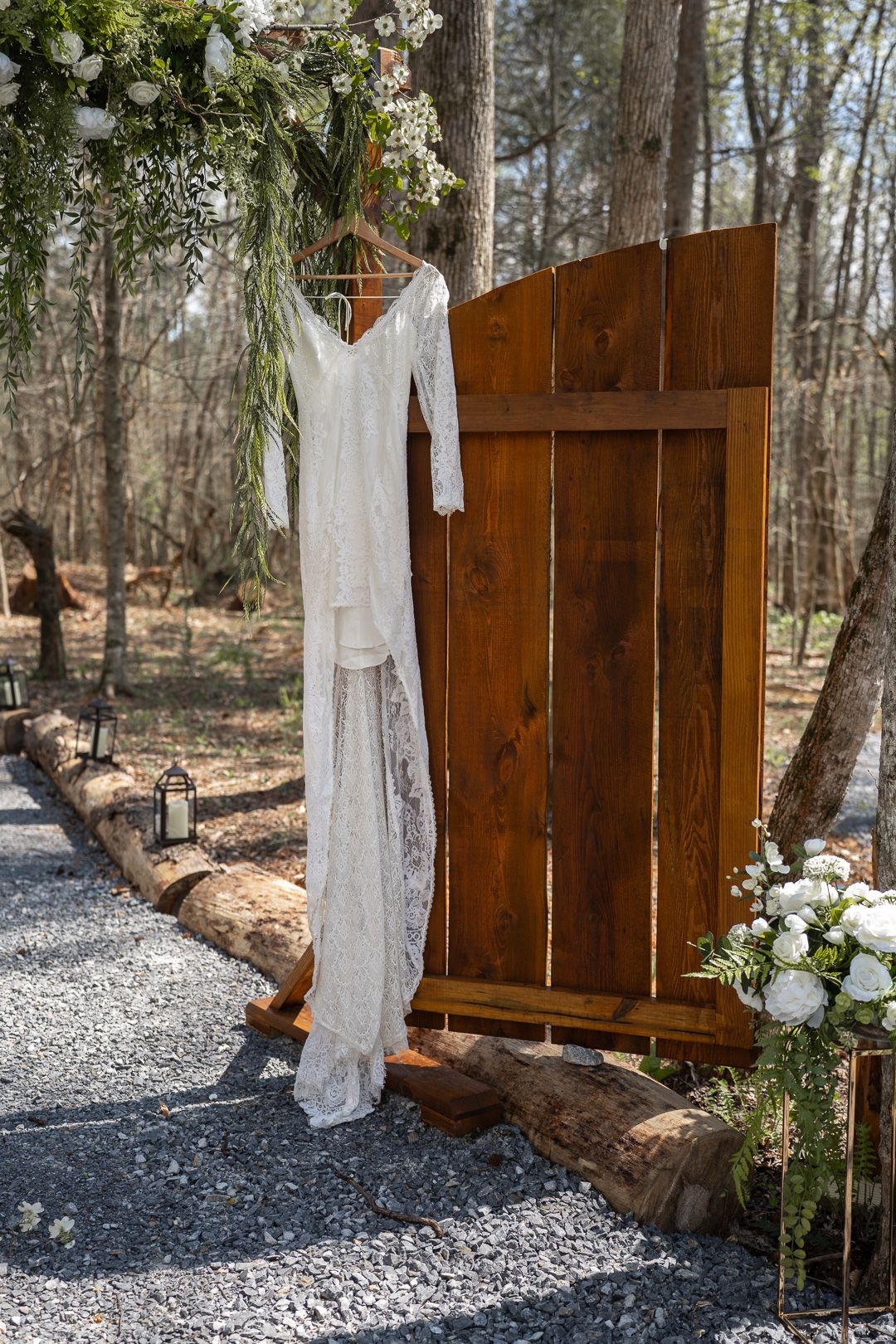 White lace wedding dress hanging on rustic wooden door backdrop surrounded by greenery and white flowers in outdoor forest setting.