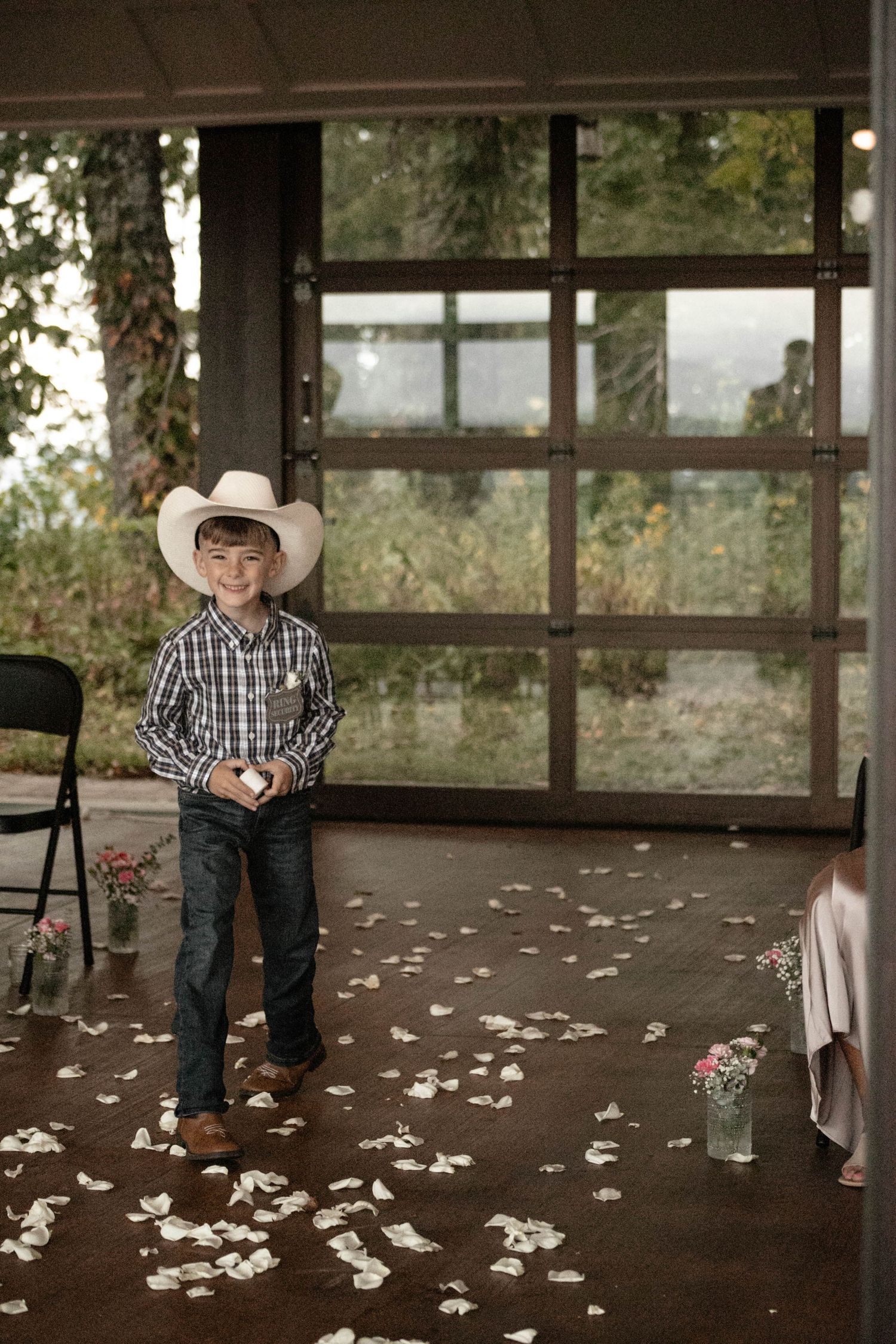 Young boy in cowboy hat and plaid shirt stands on flower petal-strewn aisle at rustic wedding venue.