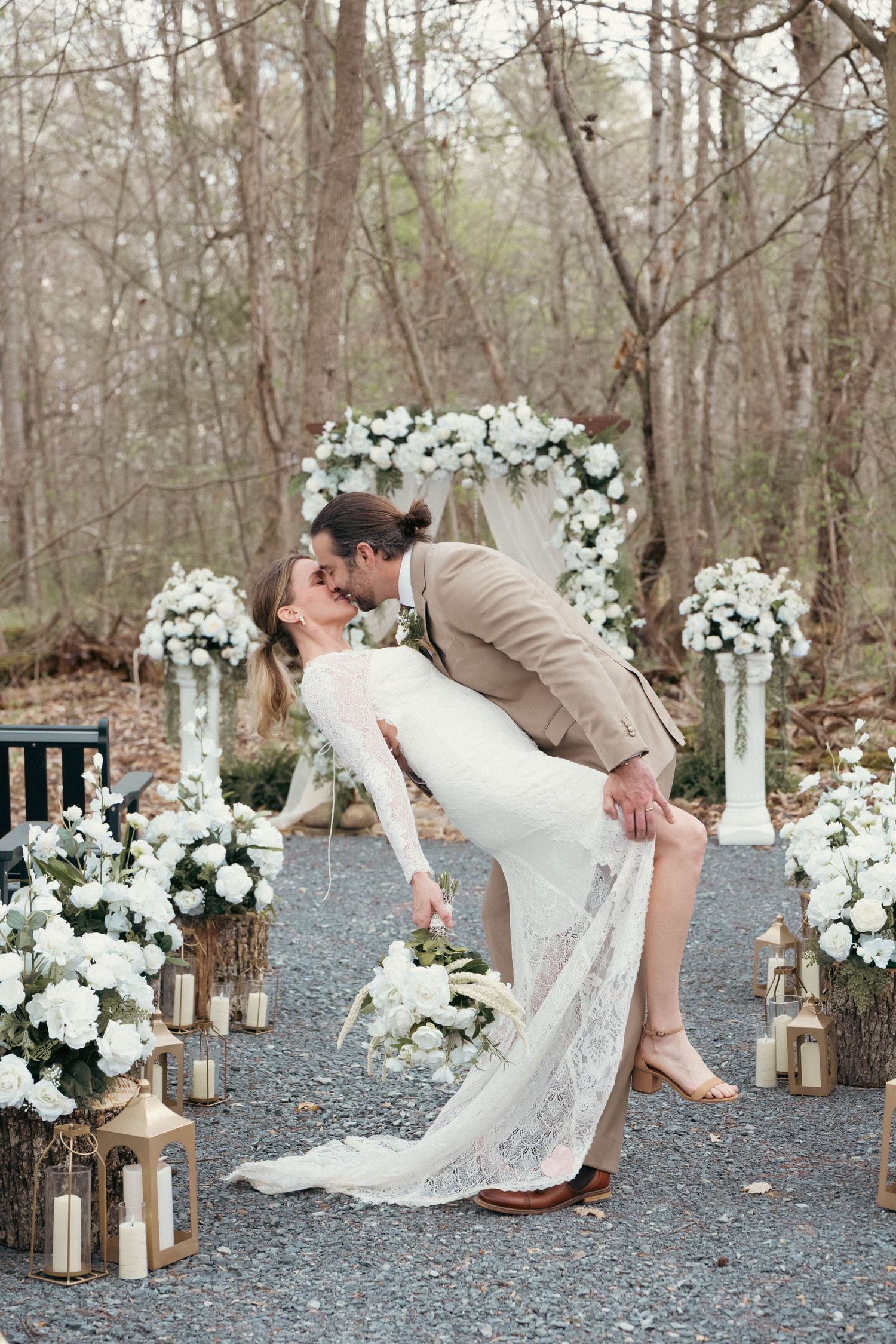Bride and groom share a romantic dip kiss at an outdoor wedding ceremony with white floral arch and lanterns.
