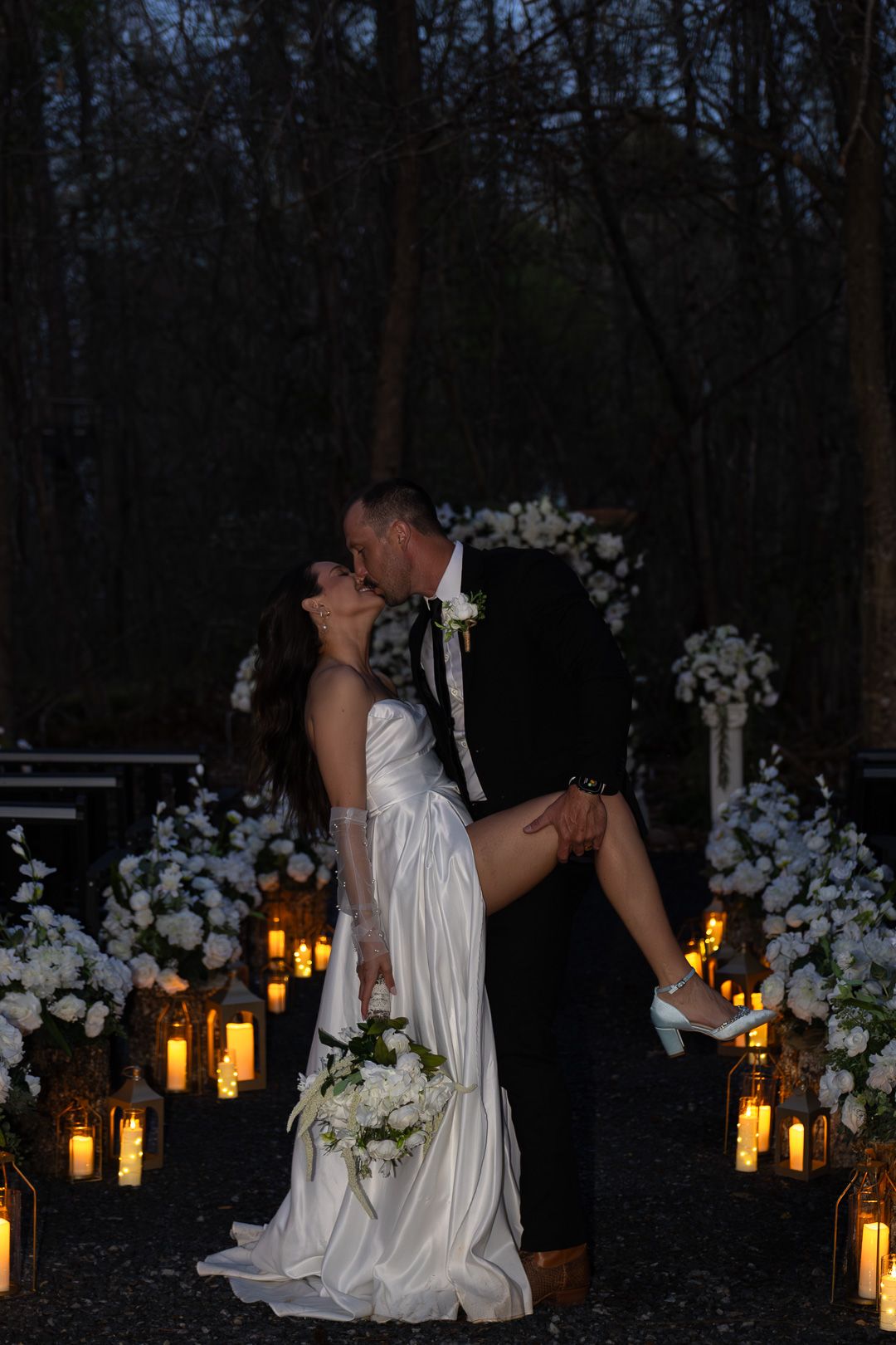 Bride and groom sharing a romantic dip kiss surrounded by glowing candle lanterns and white floral arrangements at night.