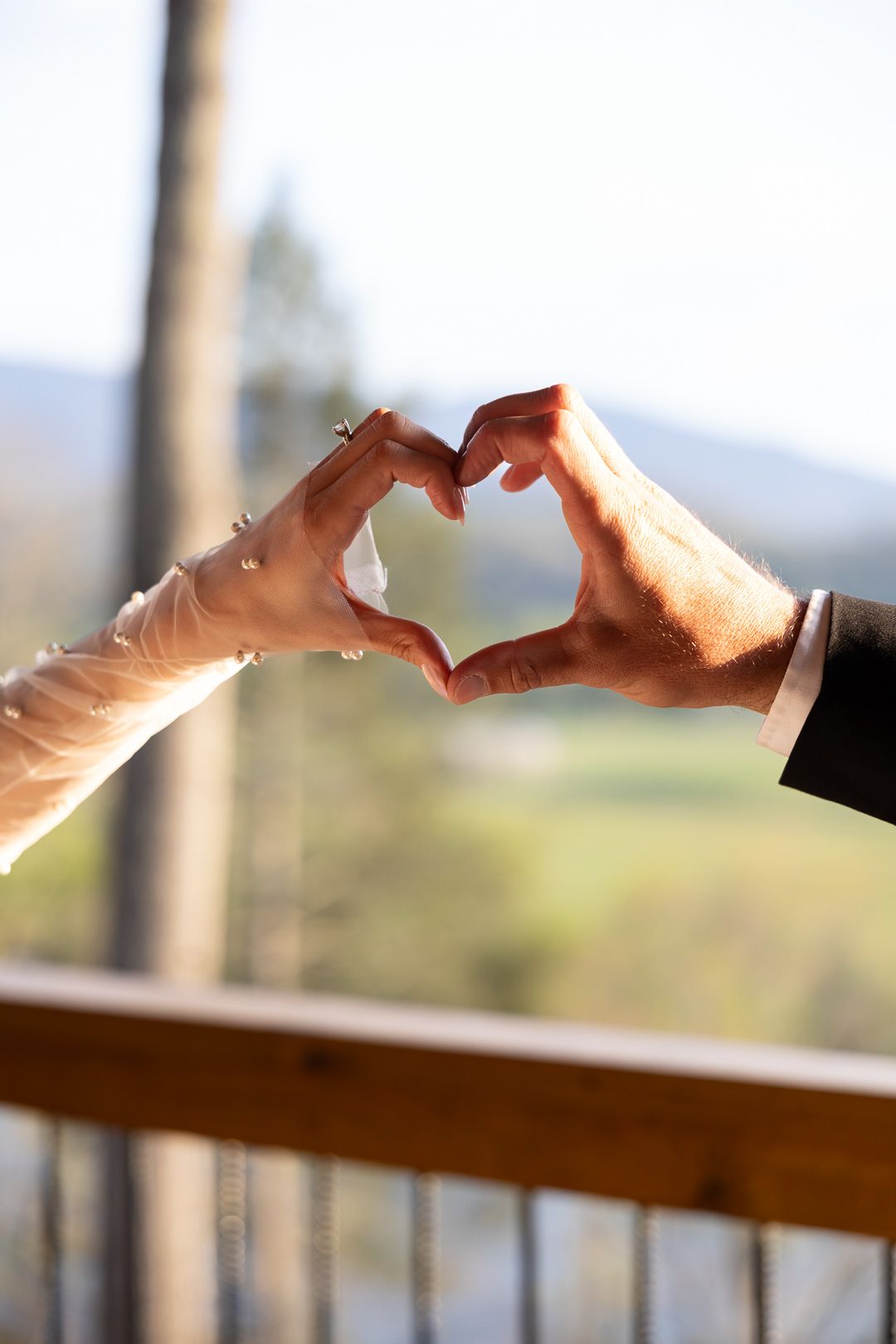 Bride and groom forming a heart shape with their hands on a sunlit wooden balcony with mountain views.