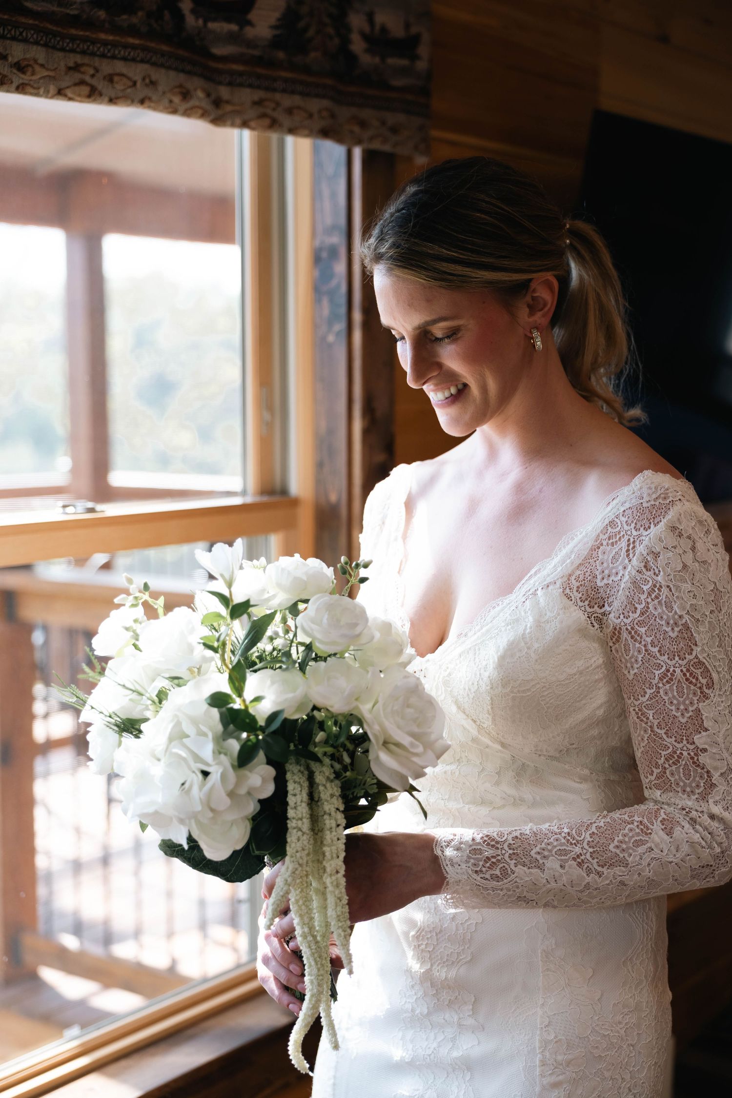 Smiling bride in lace long-sleeve wedding dress holding white floral bouquet near sunlit wooden window.