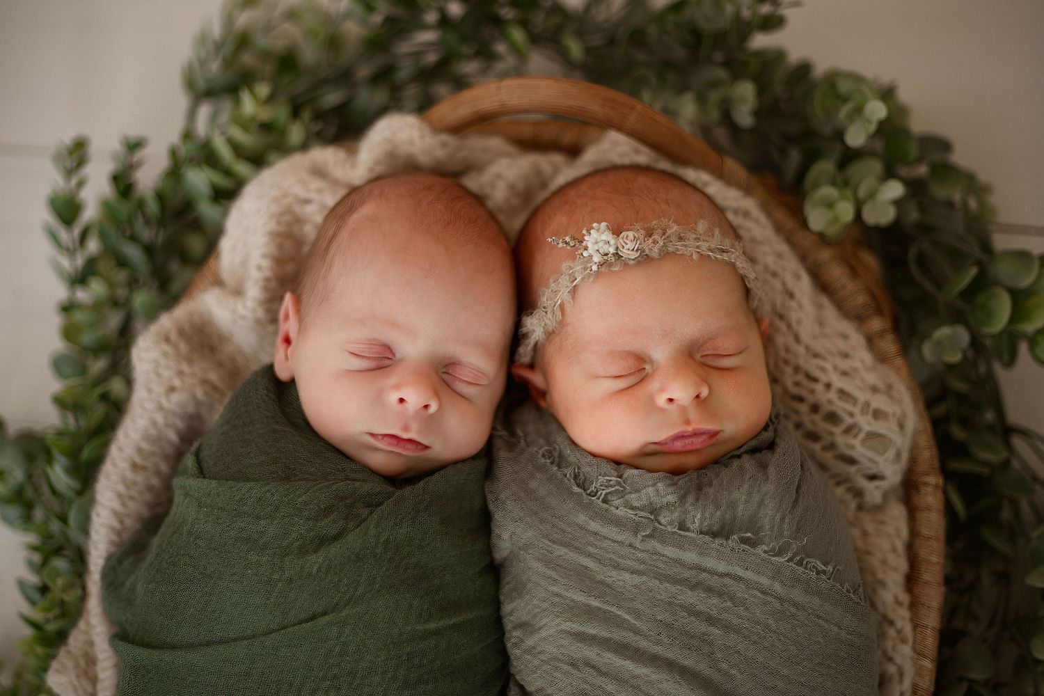 Two swaddled newborns sleeping side by side with delicate headpieces among greenery decor.