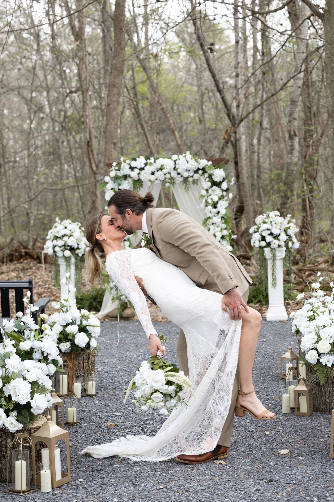 Groom dips bride for a romantic kiss at an outdoor woodland wedding ceremony surrounded by white floral arrangements.