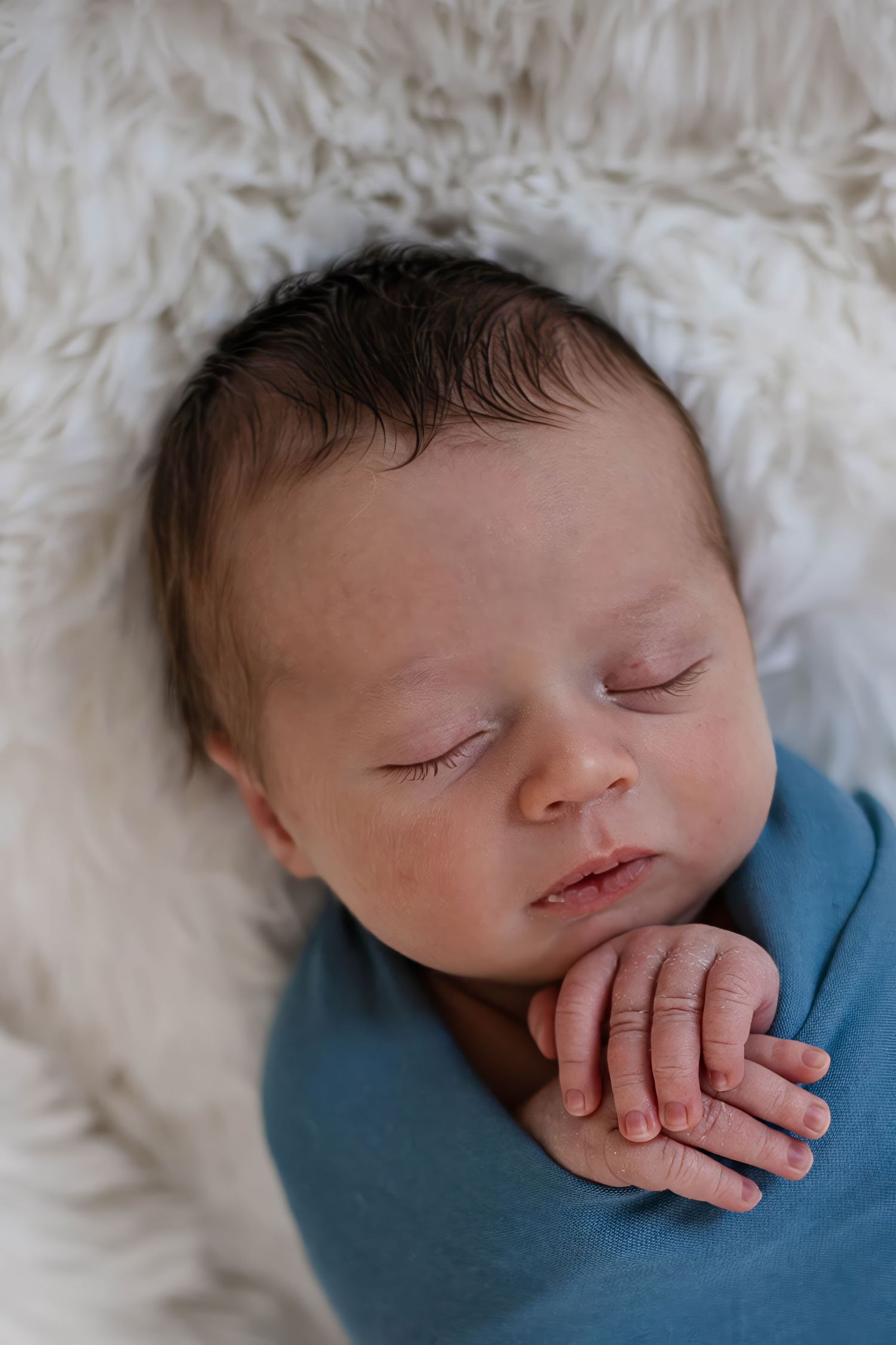 A peaceful sleeping newborn baby wrapped in a blue swaddle rests on soft white fur bedding.
