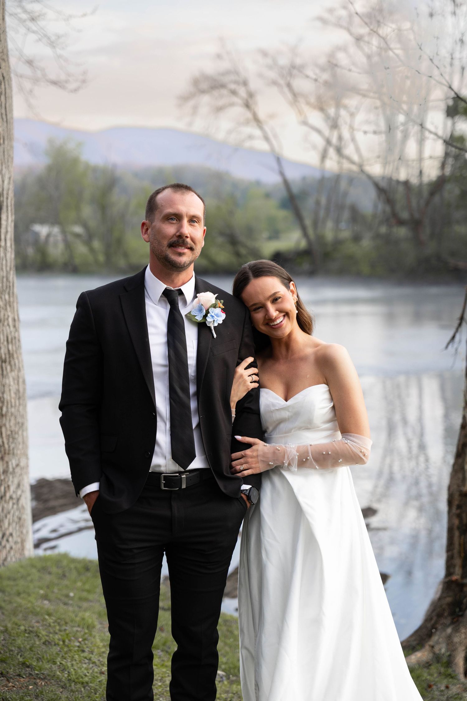 Bride and groom posing by a serene river, she in a white strapless gown, he in a black suit with boutonniere.