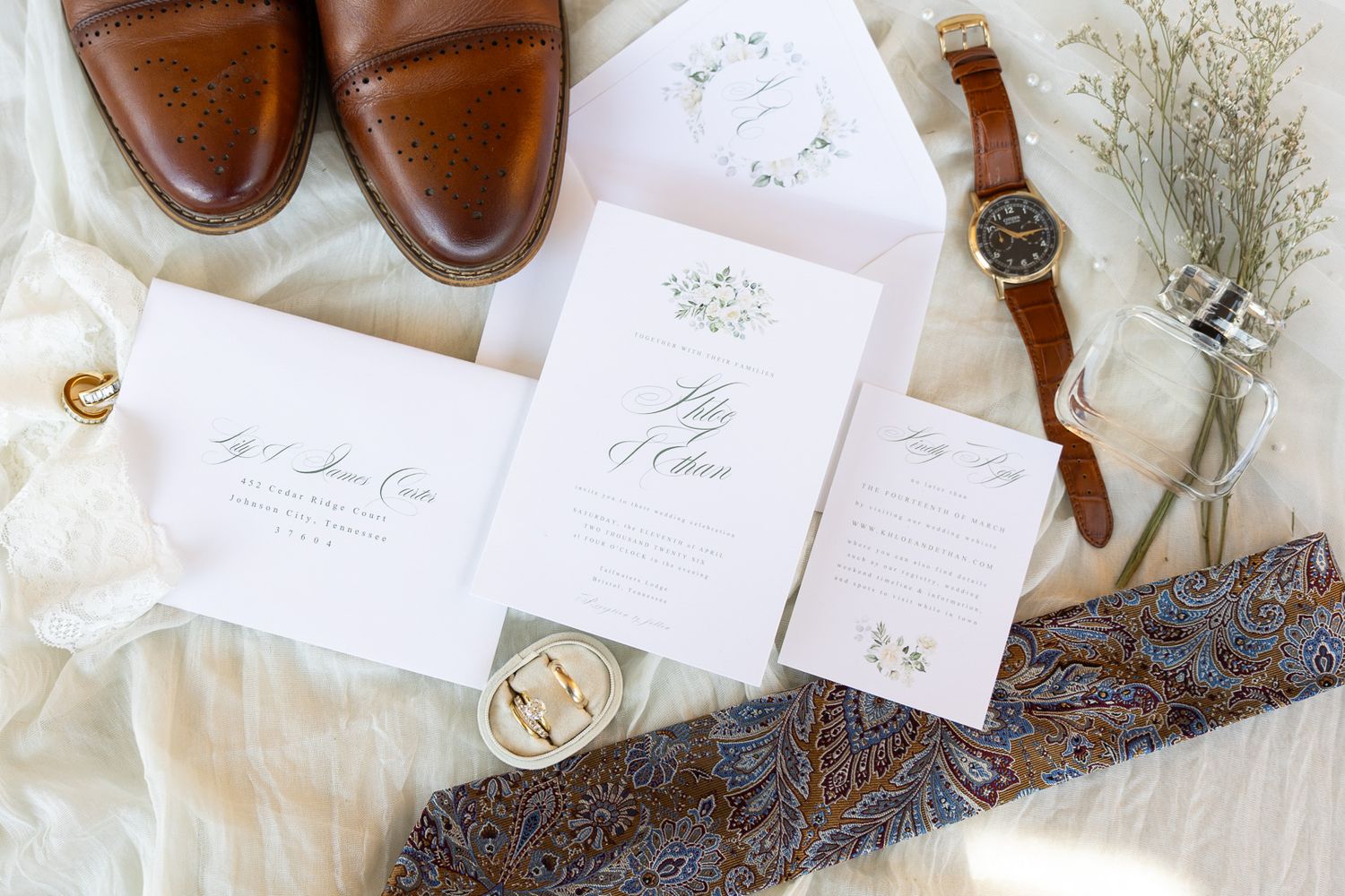 Elegant wedding flat lay with floral invitations, brown leather shoes, vintage watch, gold ring, and paisley tie on white fabric.
