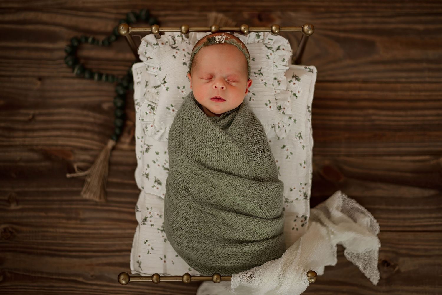 A newborn baby wrapped in a sage green swaddle blanket lies on a white lace doily against rustic wood planks.