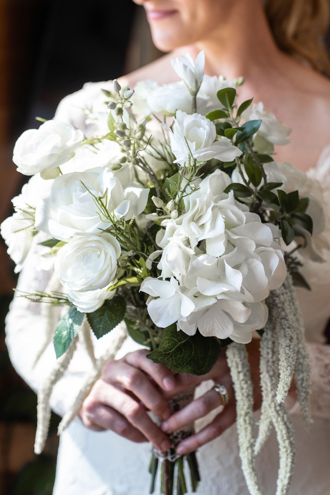 Bride holds elegant white wedding bouquet featuring hydrangeas, roses, and greenery with delicate lace ribbon accents.