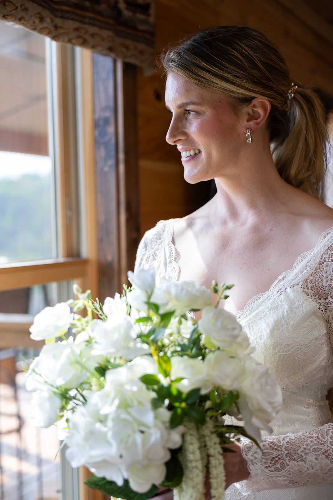 Smiling bride in lace gown holds white floral bouquet by a sunlit wooden window on her wedding day.