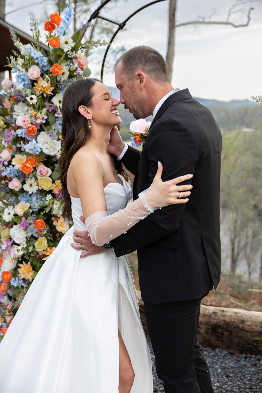 Bride and groom share a romantic embrace at floral arch wedding ceremony with colorful blooms outdoors.