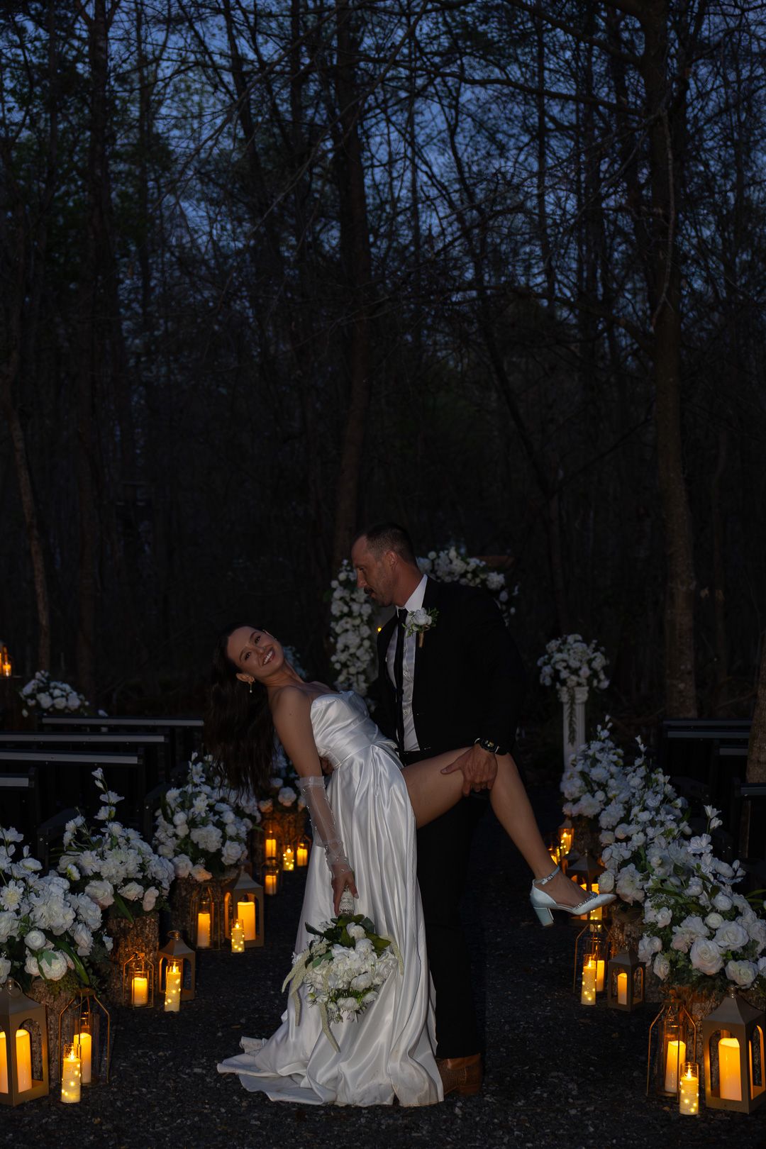 Groom dips bride among glowing lanterns and white floral arrangements in a twilight forest ceremony.