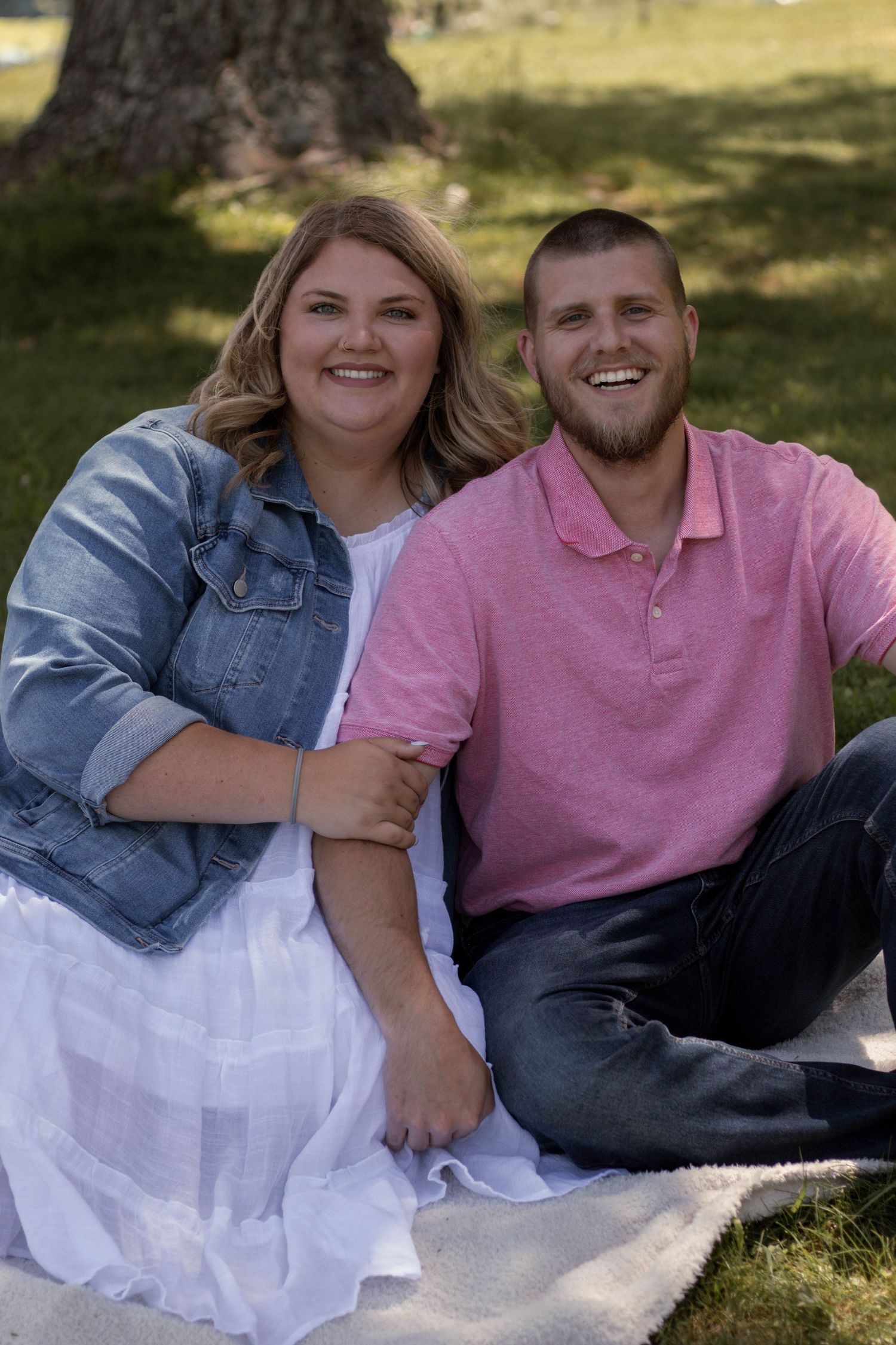 Couple sitting on blanket outdoors, wearing casual attire and sharing genuine smiles.