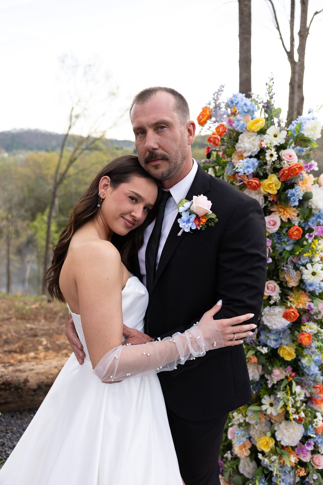 Newlyweds embrace beside a vibrant floral arch in a romantic outdoor wedding portrait.