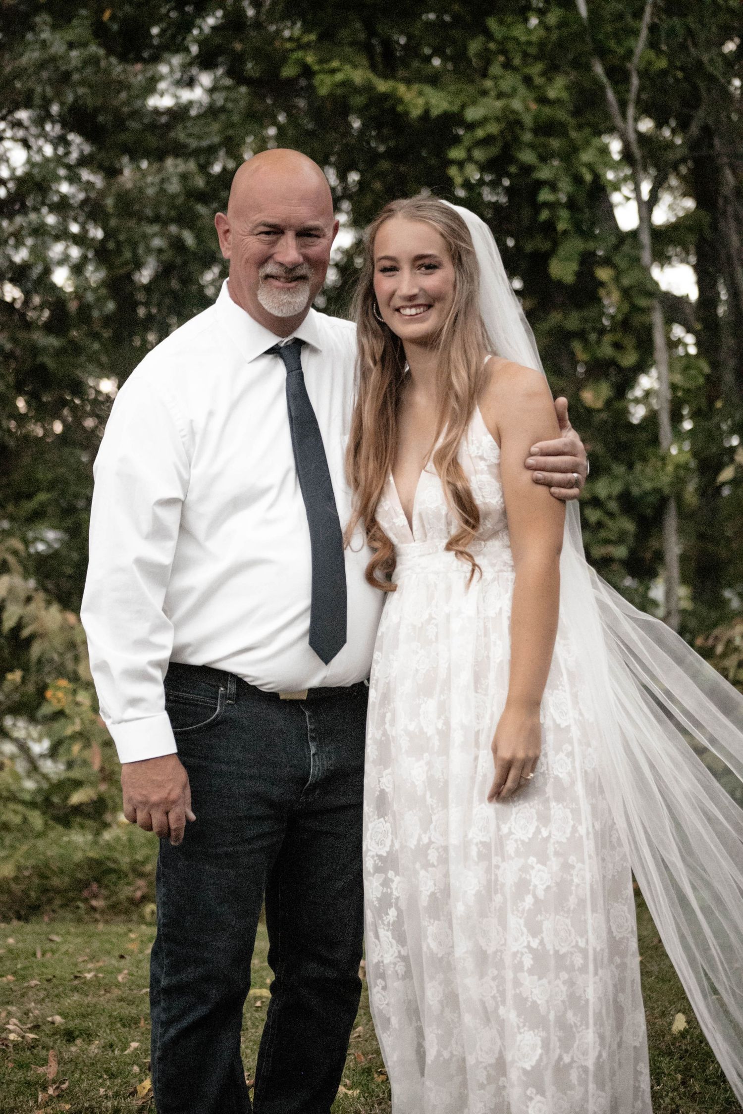 Smiling bride in lace wedding gown and veil poses with older bald man in white shirt and tie outdoors.